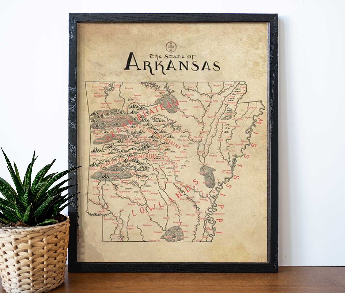 The Lord of Maps Arkansas Map is a framed, hand-drawn vintage-style print displaying rivers, mountains, and labeled neighboring states in red. It leans against a wall on wood beside a small potted plant.