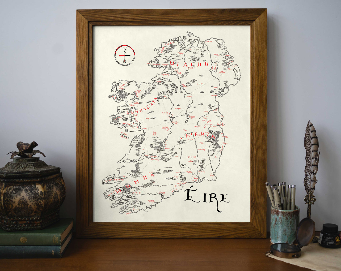 The Lord of Maps Ireland Map is a high-quality framed print featuring a hand-drawn map with Irish place names, beautifully displayed on a wooden desk alongside books, pens, a feather quill, and a decorative container.