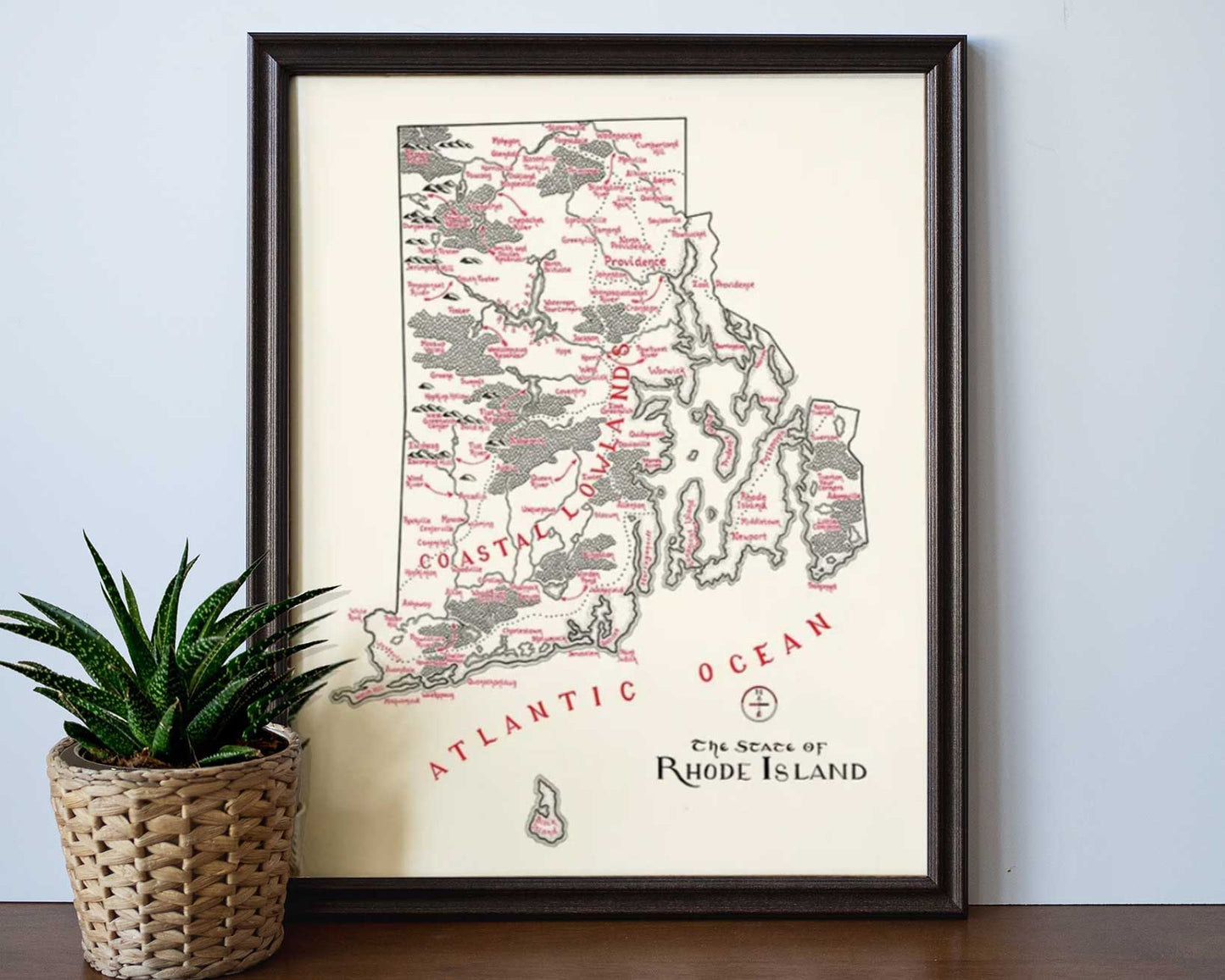 The Lord of Maps Rhode Island Map is a framed hand-drawn print with red-labeled towns and coasts on a light background, shown on a wooden surface beside a small potted plant—perfect wall art for any space.
