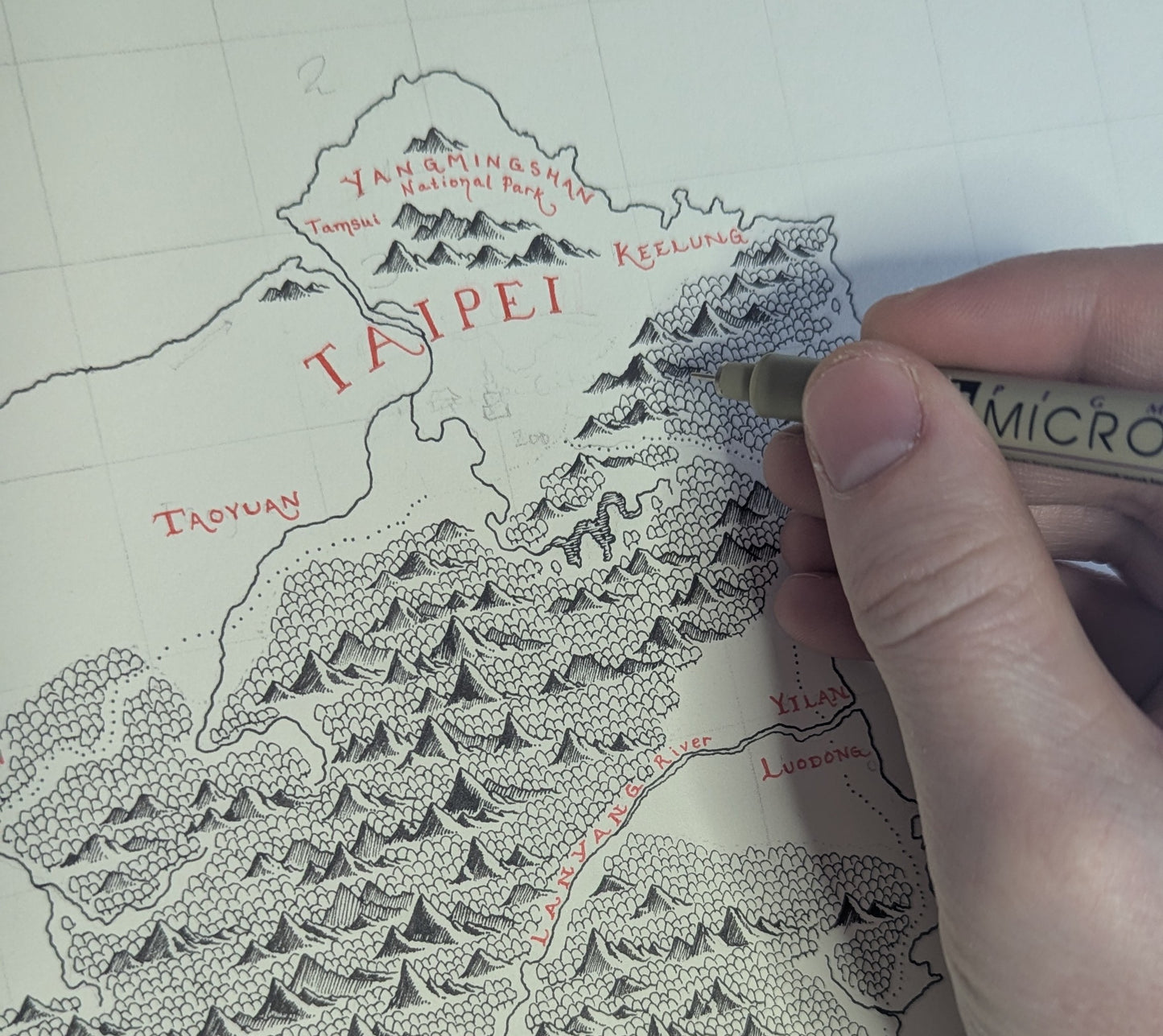 A hand uses a fine-tip Micron pen to draw detailed mountains on the Taiwan Map by Lord of Maps, labeling spots like Taipei, Taoyuan, Keelung, and Yangmingshan National Park in red ink—ideal for framing or as a high-quality print.