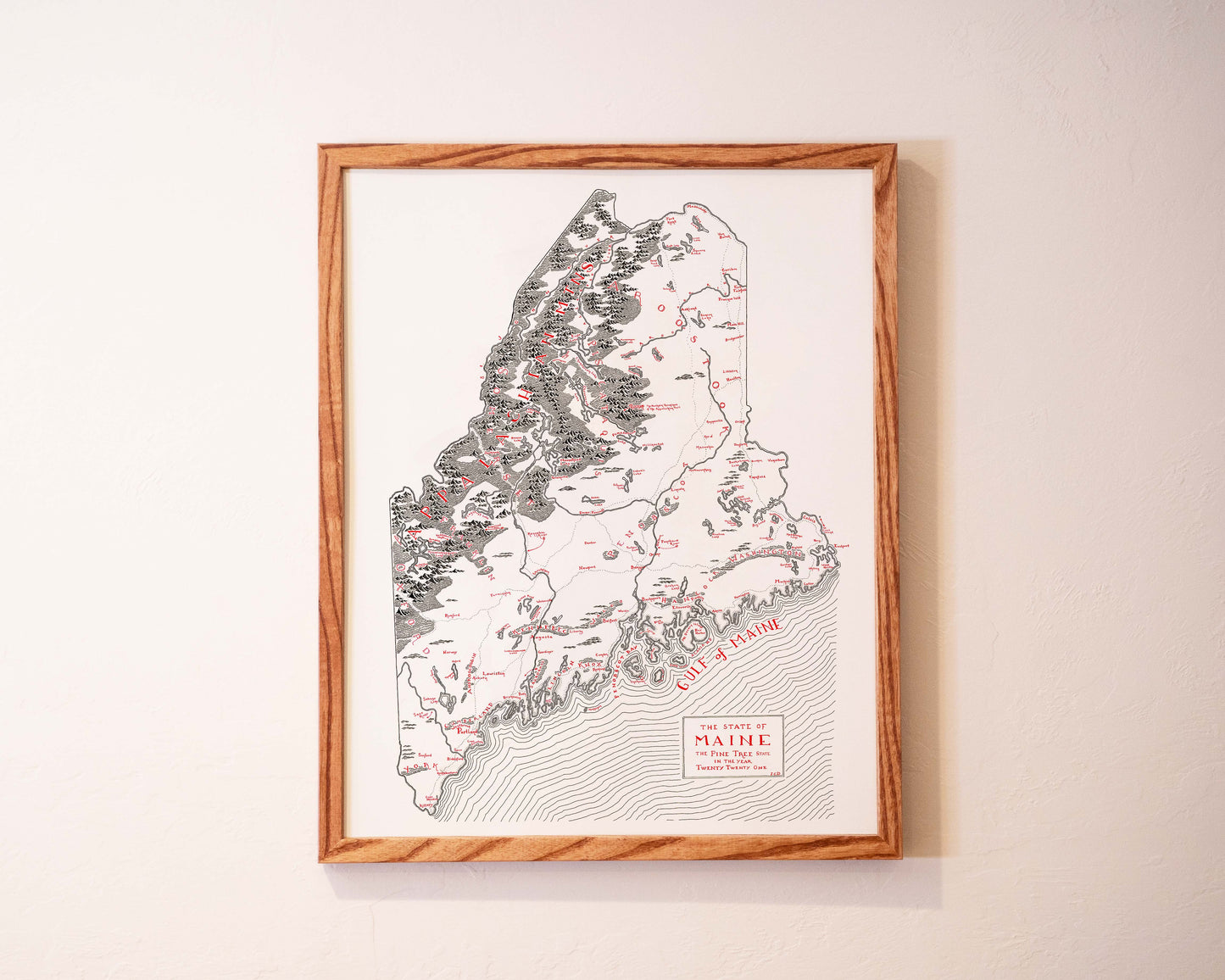 Framed black-and-white illustrated map of the state of Maine, showing detailed topography and labeled locations, hung on a light-colored wall.