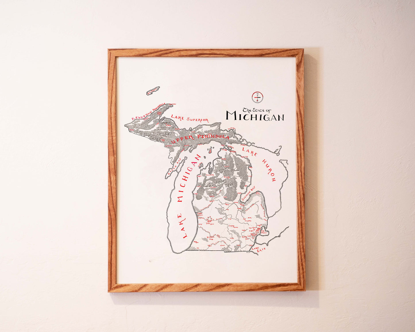 A framed hand-drawn map of Michigan hangs on a white wall, showing the state’s outline, lakes, and key locations labeled in black and red ink.