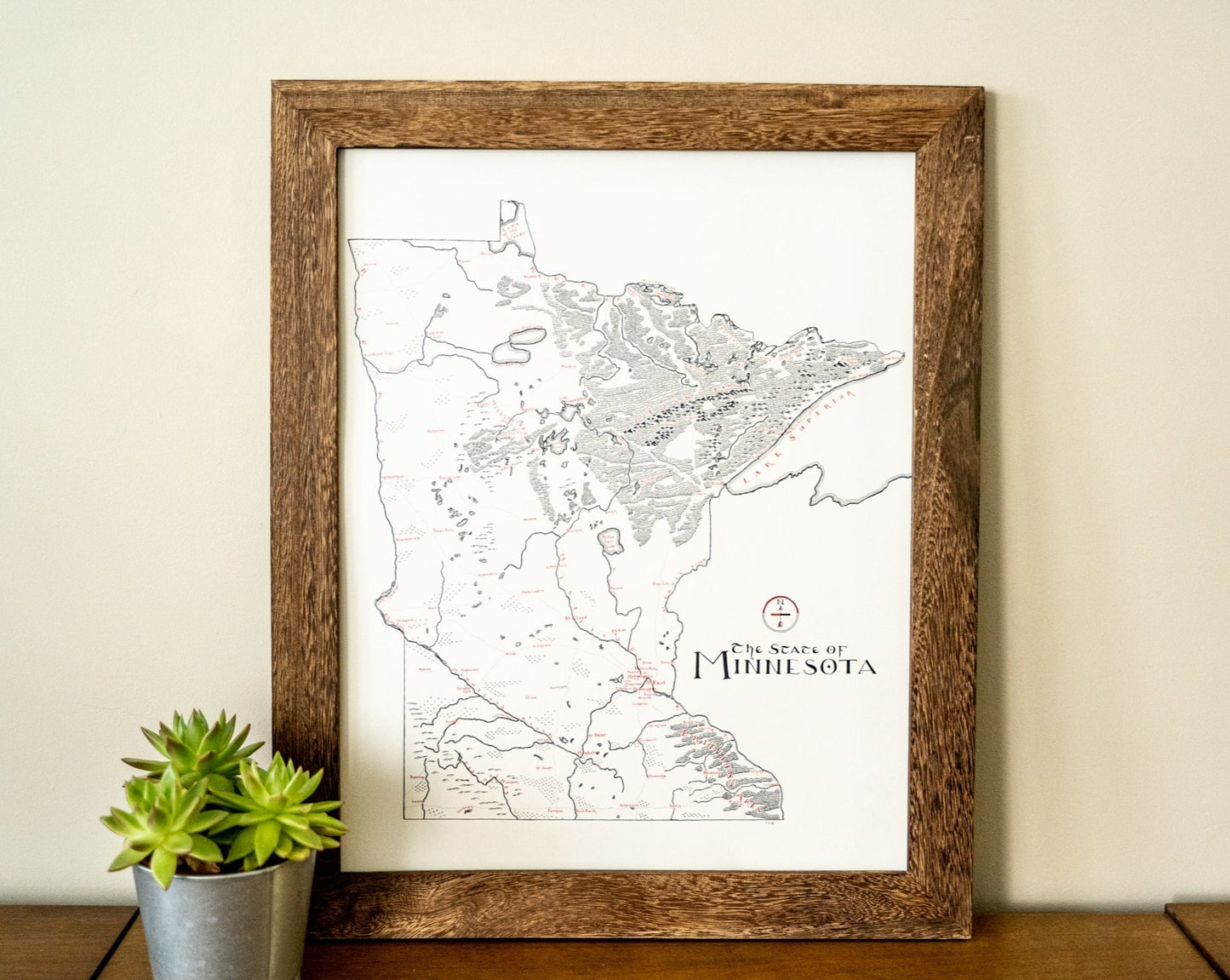 The Minnesota Map by Lord of Maps hangs framed on a beige wall, while a small green succulent in a gray pot rests on the wooden surface below this detailed hand-drawn map print.