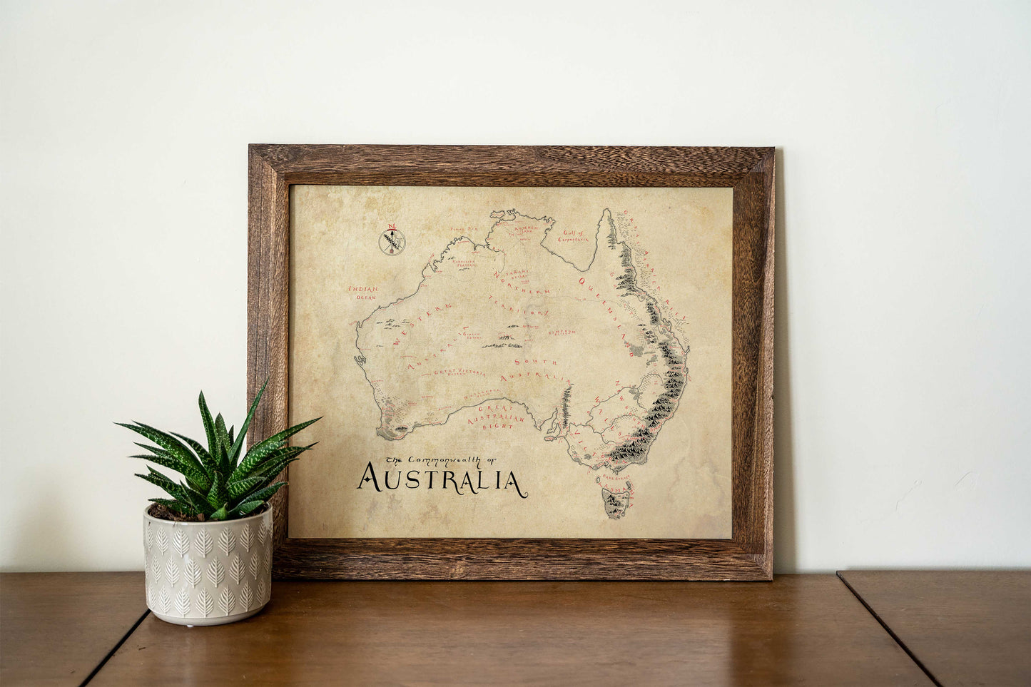 The Lord of Maps Australia Map, a hand-drawn print in an elegant wooden frame, is displayed on a wooden surface beside a potted succulent against a plain white wall.
