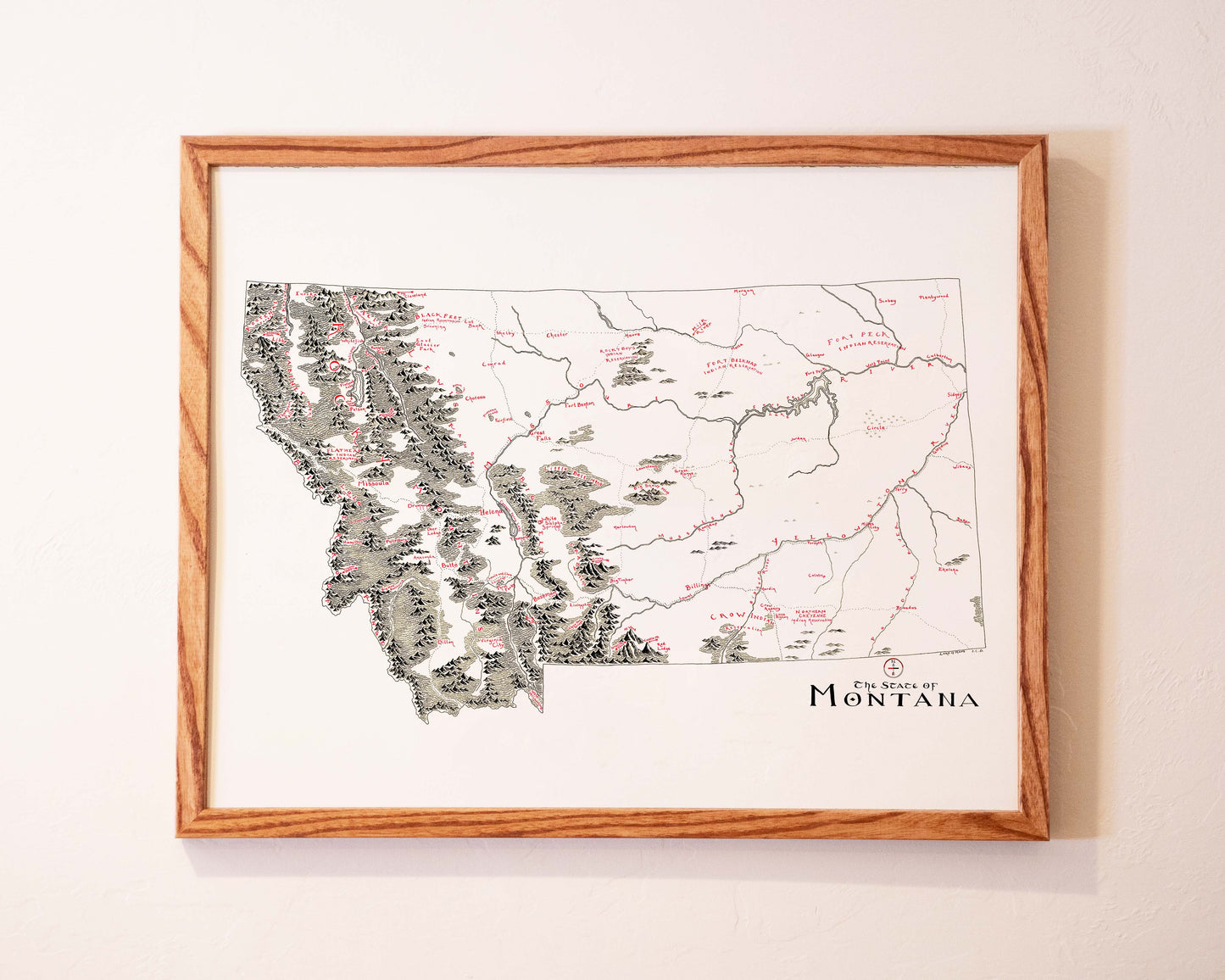 A framed map of Montana hangs on a light-colored wall, showing state borders, major cities, rivers, and mountain ranges with labeled geographic features in black and red text.