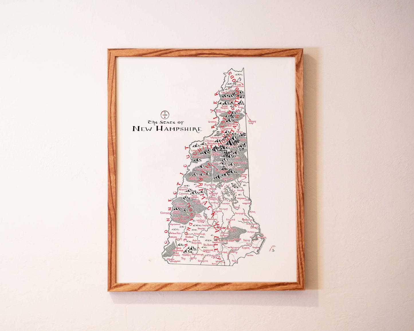 Framed vintage-style map of New Hampshire with black and red text and markings, hanging on a light-colored wall.