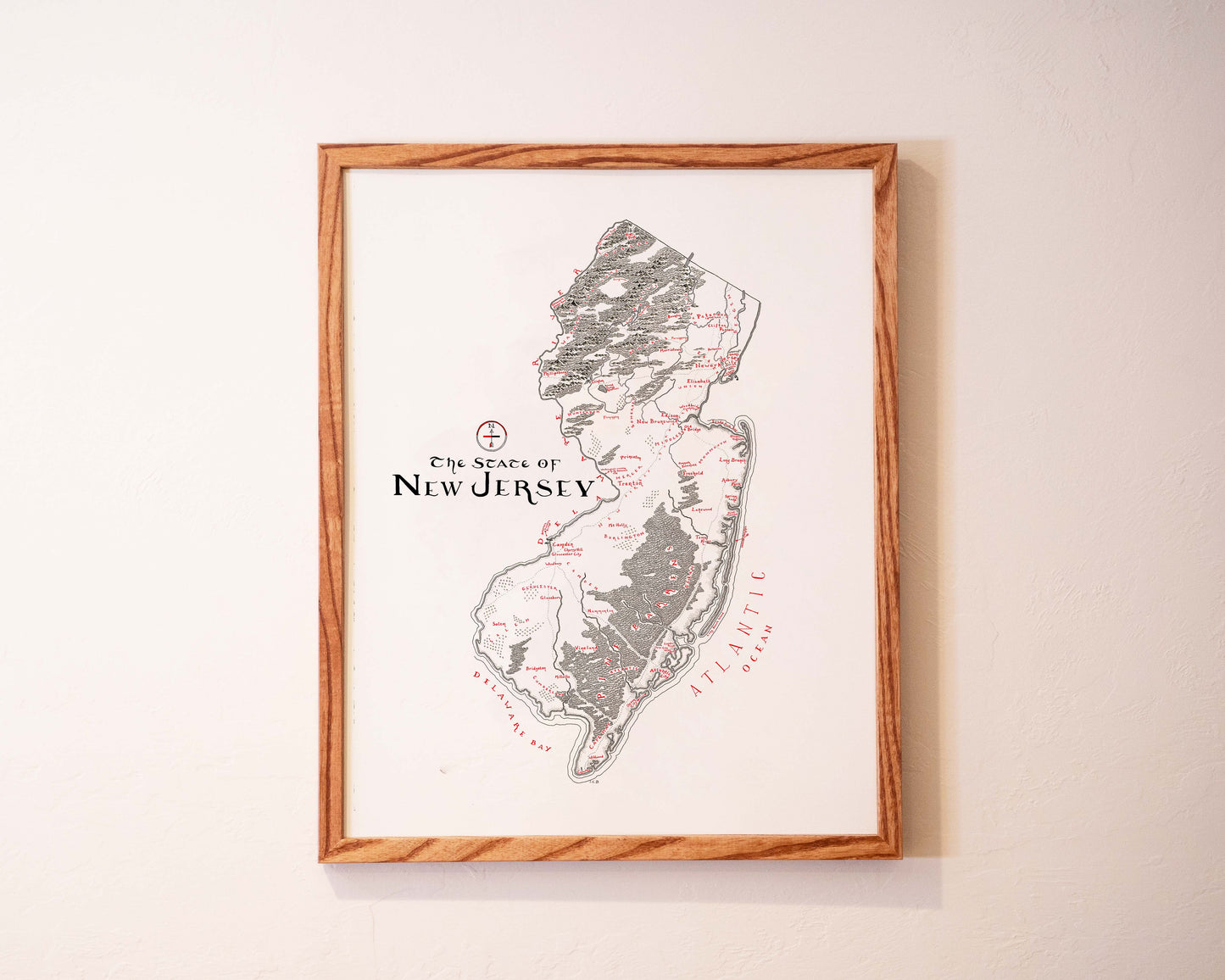 A framed map of New Jersey hangs on a light-colored wall. The map is labeled “The State of New Jersey” and shows geographic features and some city names, with ATLANTIC OCEAN written along the coast.
