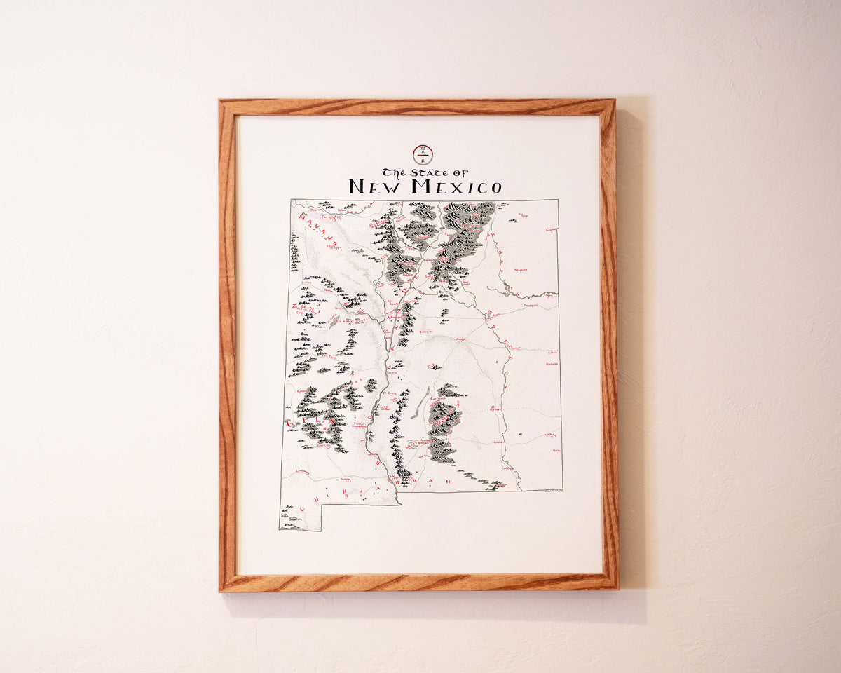 A framed map of New Mexico with black and red markings hangs on a light-colored wall. The title The State of New Mexico is written at the top of the map in decorative font.
