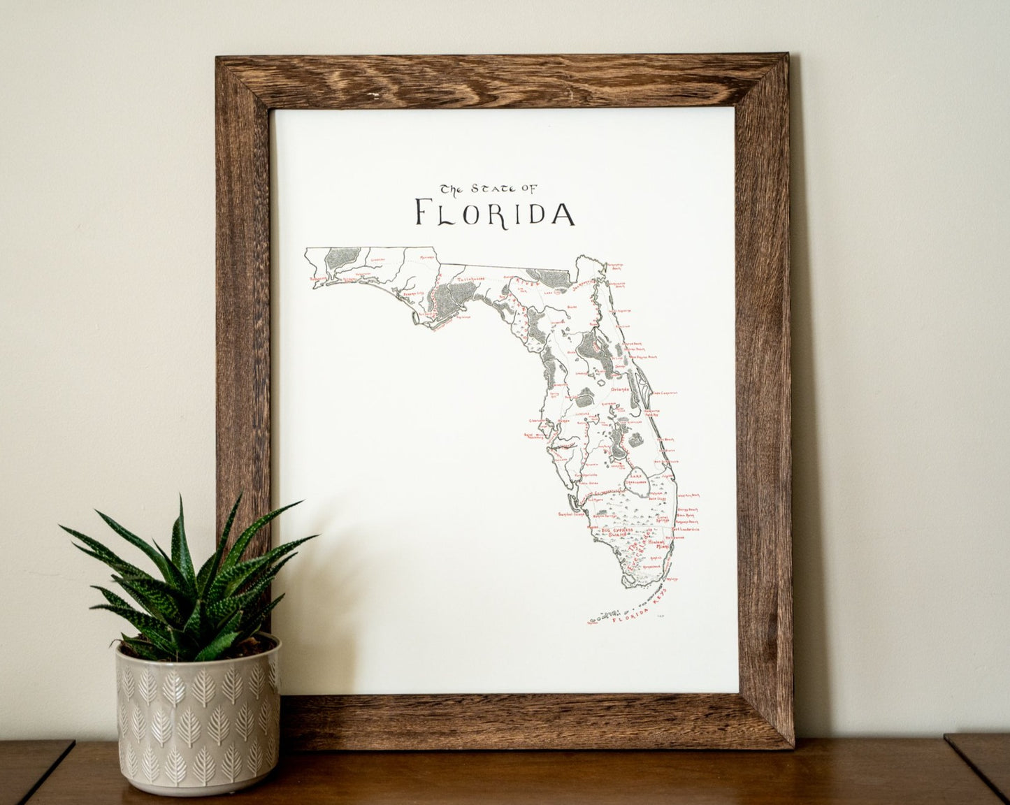 The Lord of Maps Florida Map is a framed, hand-drawn print with unique handwritten details. Displayed stylishly beside a potted succulent, it makes perfect wall art for any space.