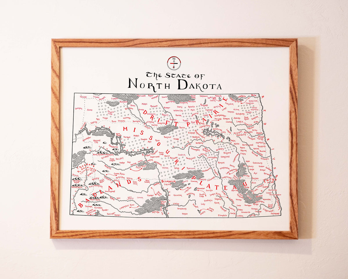 Framed map of North Dakota showing rivers, towns, and regions in black, with Native American tribal names labeled in red across the state. The map title is written at the top in stylized text.