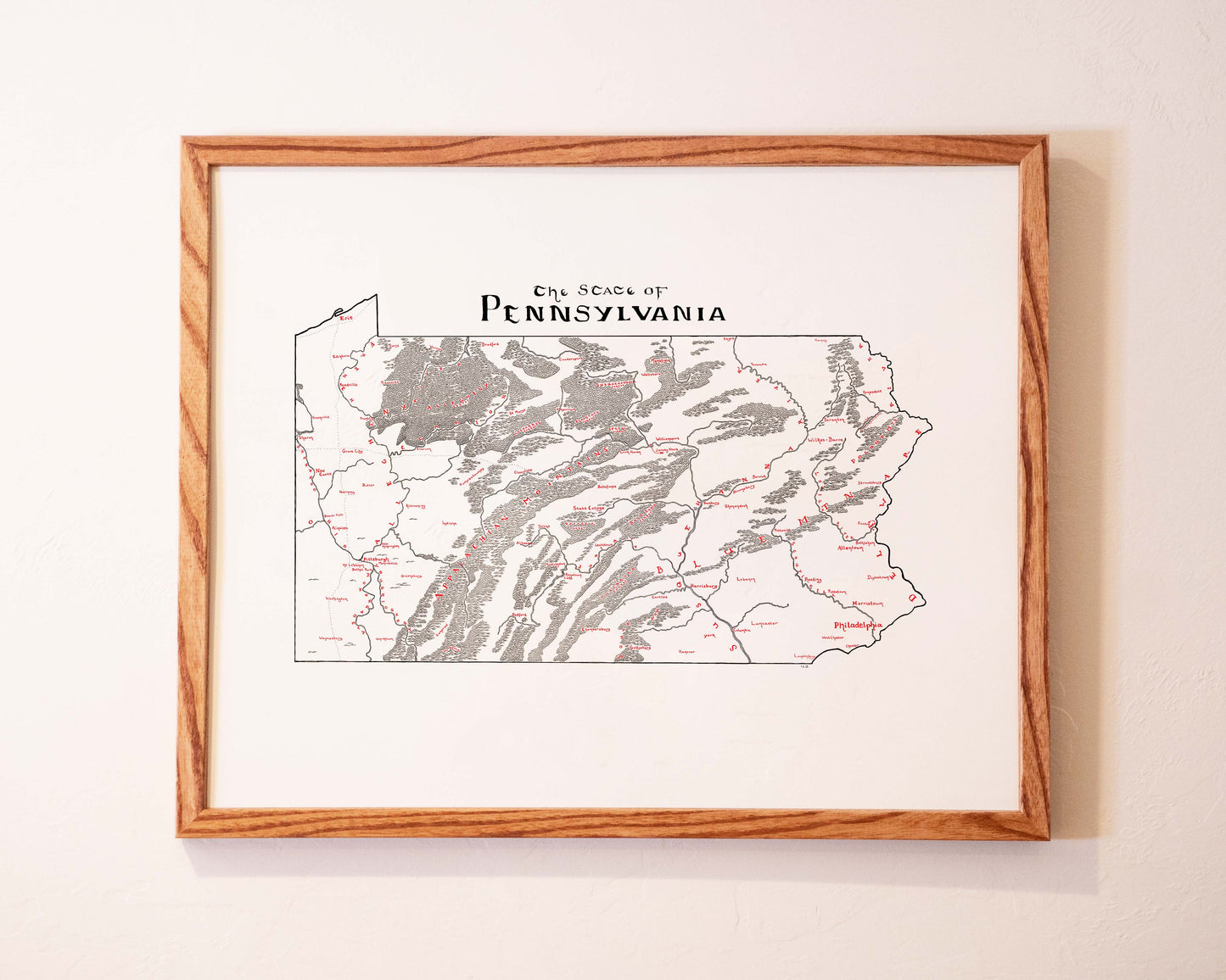 A framed map of Pennsylvania hangs on a white wall. The map outlines the state, highlighting cities, rivers, and mountain ranges, with The State of Pennsylvania written at the top.