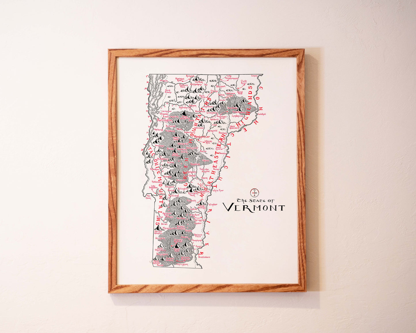 Framed illustrated map of Vermont hangs on a light-colored wall, showing mountains, rivers, and towns in black and red sketch style, with the words The State of Vermont written near the bottom right.
