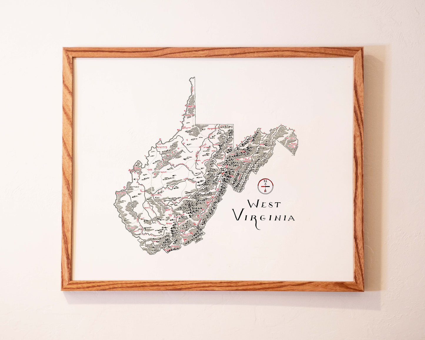 Framed map of West Virginia with detailed topographical markings and place names in black and red, hanging on a white wall. The words West Virginia are written in script on the right side.