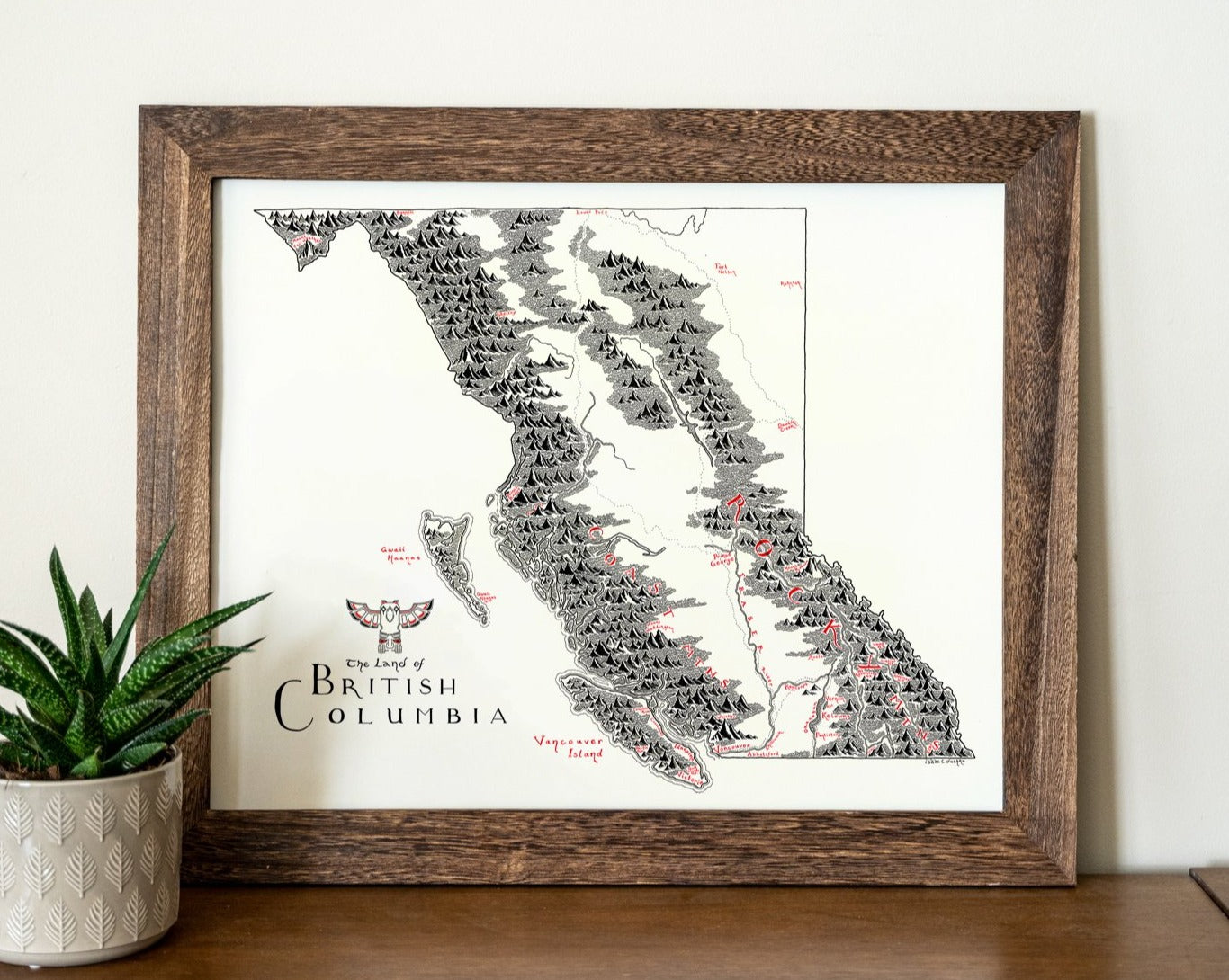 The British Columbia Map by Lord of Maps is a framed print featuring hand-drawn black and grey mountains, red place names, and a decorative title. Displayed as wall art, it rests on wood beside a potted succulent.