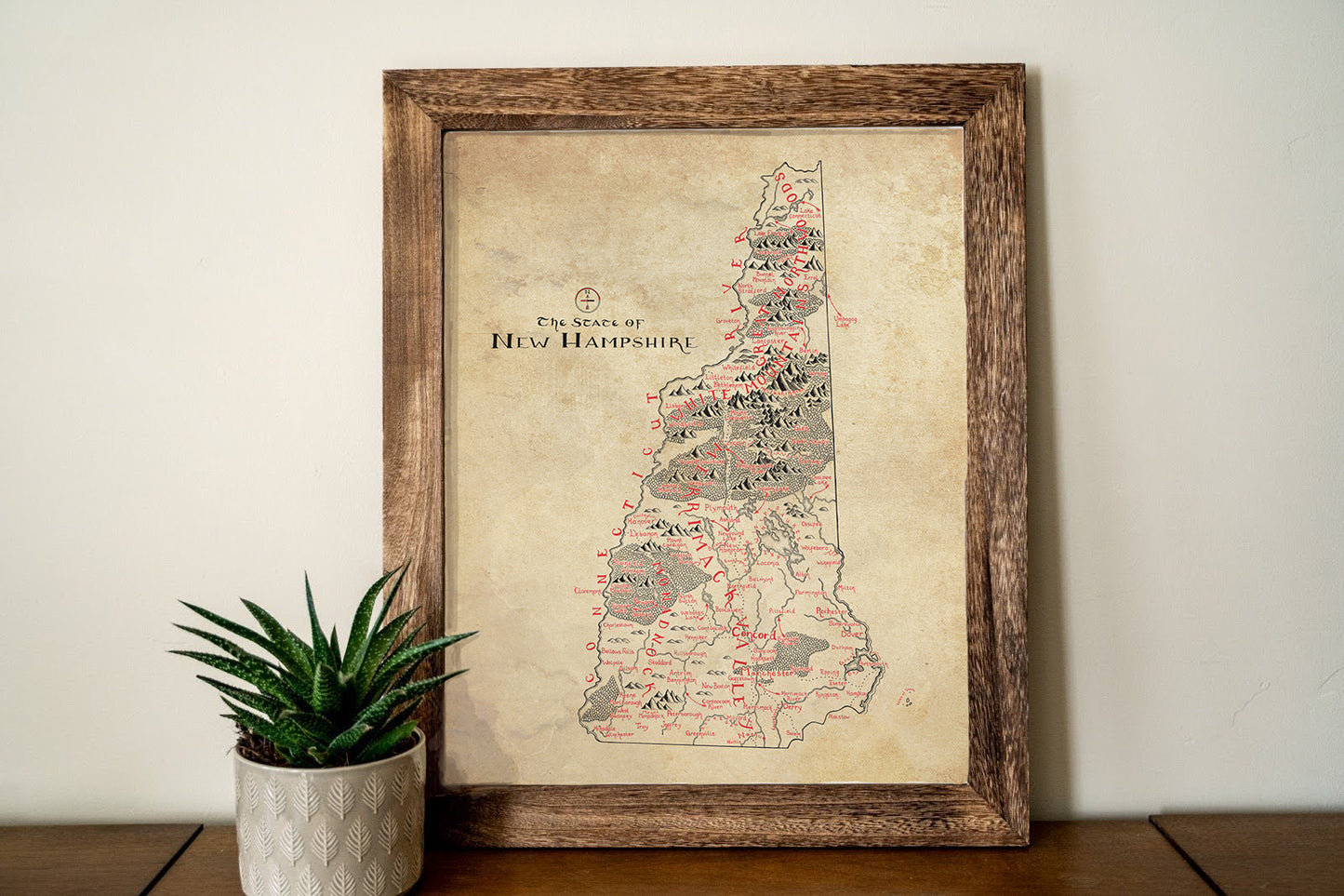 A framed Lord of Maps New Hampshire Map hangs on a light wall, with a small succulent in a patterned white pot resting on the wooden surface beneath it.