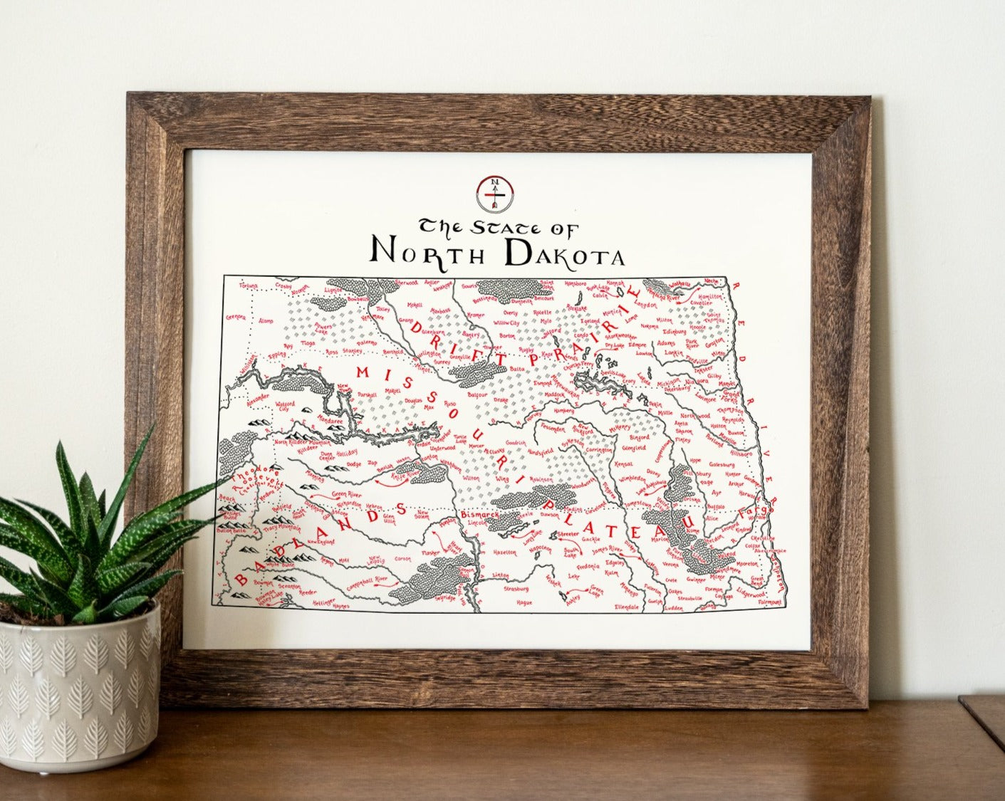 A North Dakota Map print by Lord of Maps with black and red details rests on a wooden surface beside a small potted succulent. The hand-drawn map features the label "The State of North Dakota.
