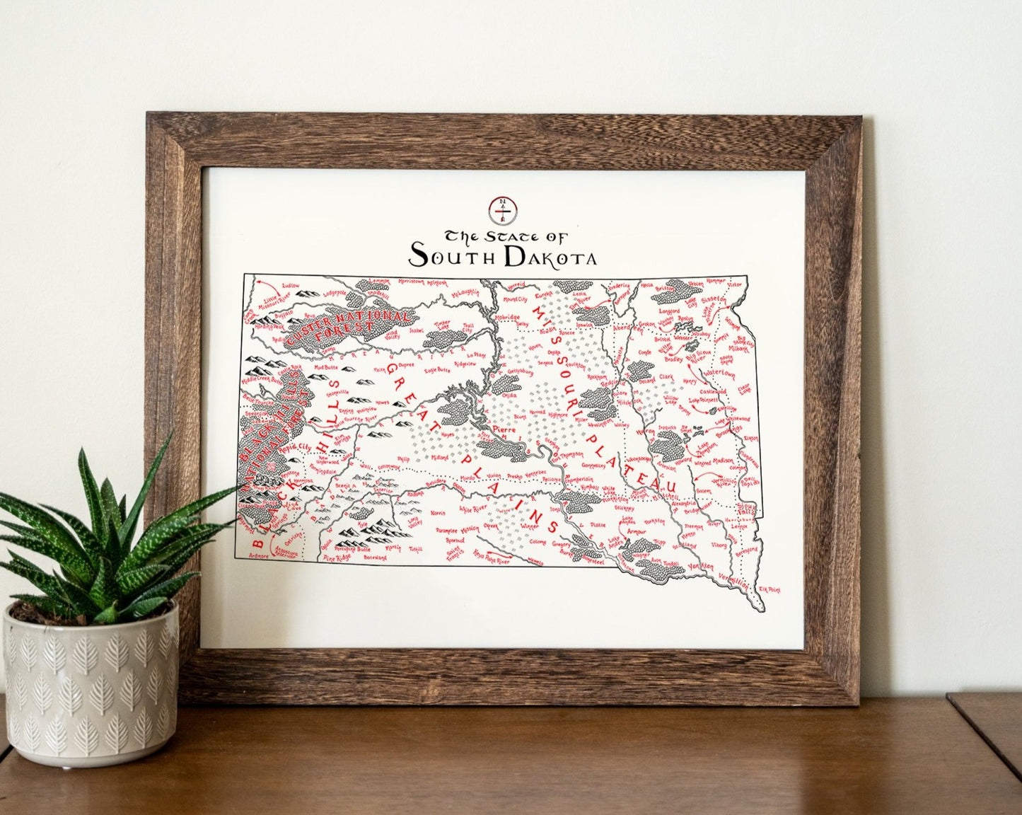 The Lord of Maps South Dakota Map is a framed wall art featuring hand-drawn landmarks and place names in black and red, shown on a wooden surface beside a potted plant.
