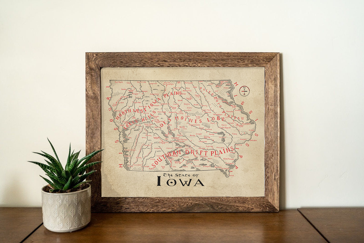 The Lord of Maps Iowa Map is a vintage-style framed print, featuring hand-drawn, labeled regions and red rivers. Displayed on wood beside a potted succulent against a white wall, it's classic map wall art for any space.