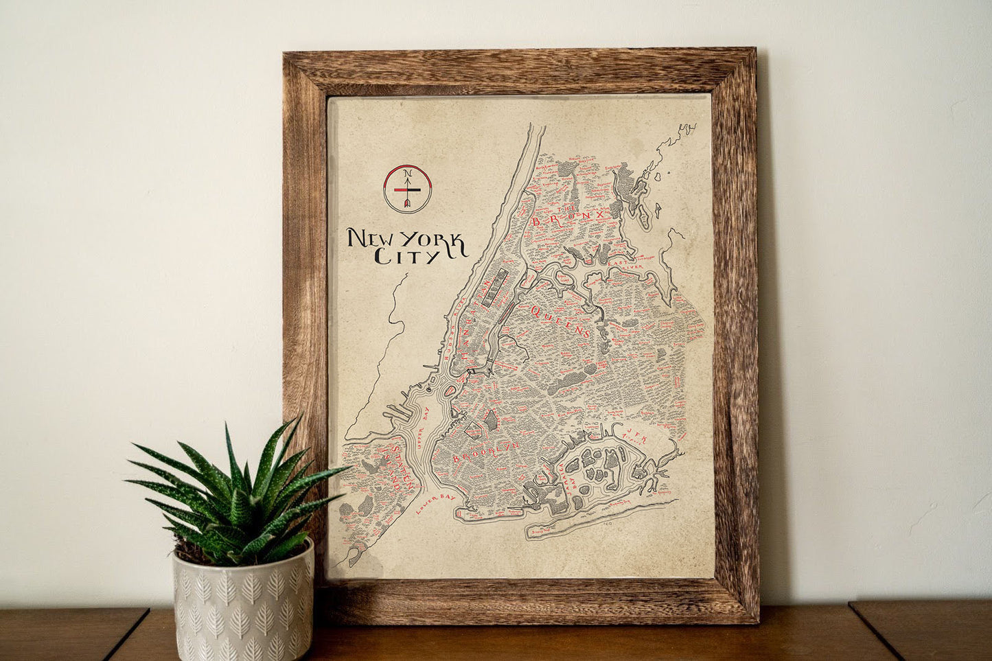 The Lord of Maps New York City Map, a framed vintage-style print with intricate hand-drawn details and bold red highlights, sits on a wooden surface beside a small succulent in a textured white pot.