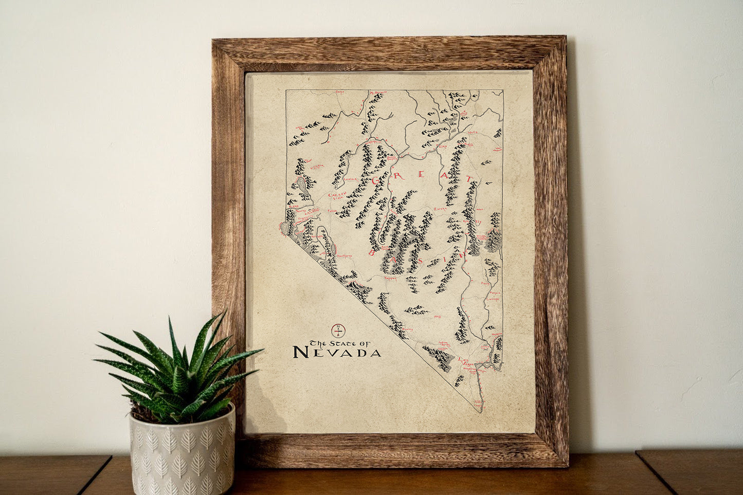 The Nevada Map by Lord of Maps features hand-drawn details and stylish text in an art frame, displayed on a wooden surface with a small potted succulent to the left.