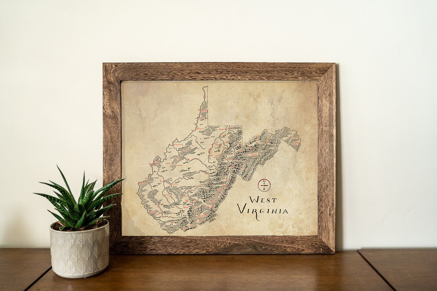 The Lord of Maps West Virginia Map features a vintage-style hand-drawn print in a frame, shown on a wooden surface beside a spiky potted plant—a perfect wall art piece for any room.