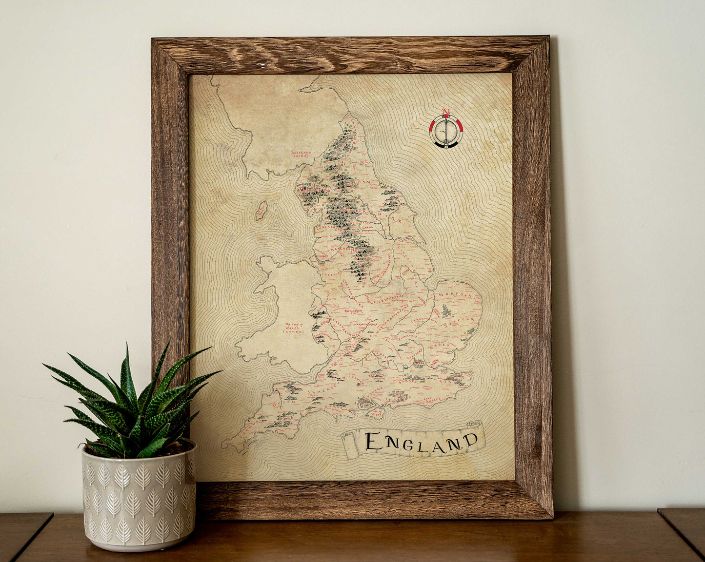 The Lord of Maps "England Map," a framed vintage-style print with classic fonts and illustrations, hangs on a light wall. Next to it, a patterned white pot holds a succulent on the wooden surface.