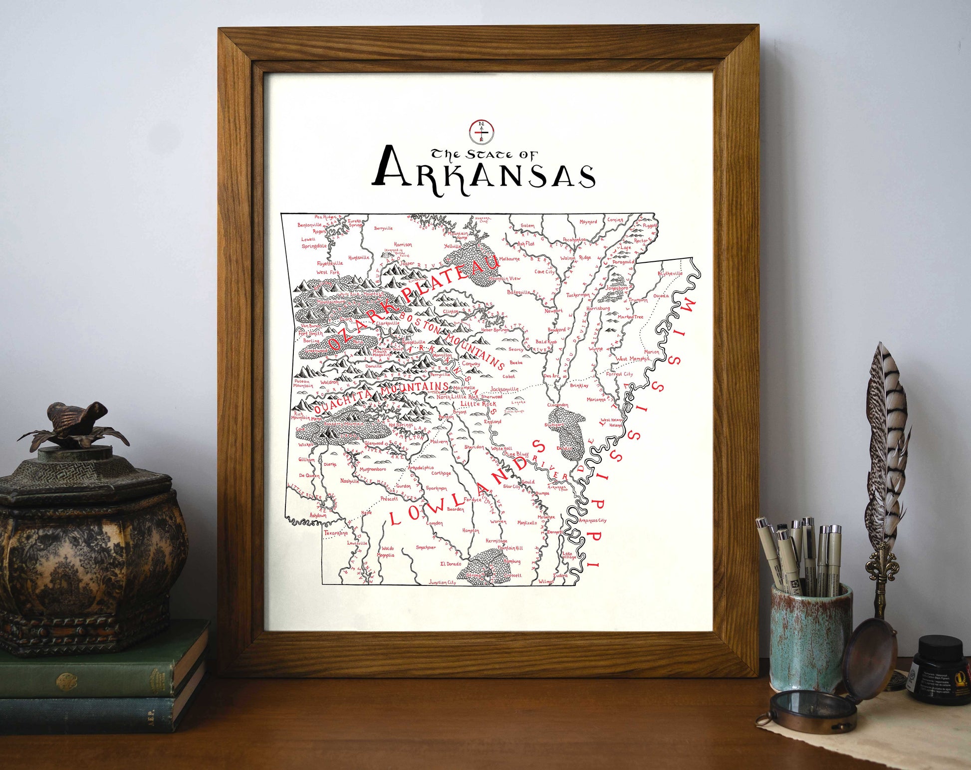 The Lord of Maps Arkansas Map is a framed, vintage-style print showing the state’s counties and rivers with “ARKANSAS” at the top. It’s displayed on a desk beside books, a cup with pens, and a decorative container.