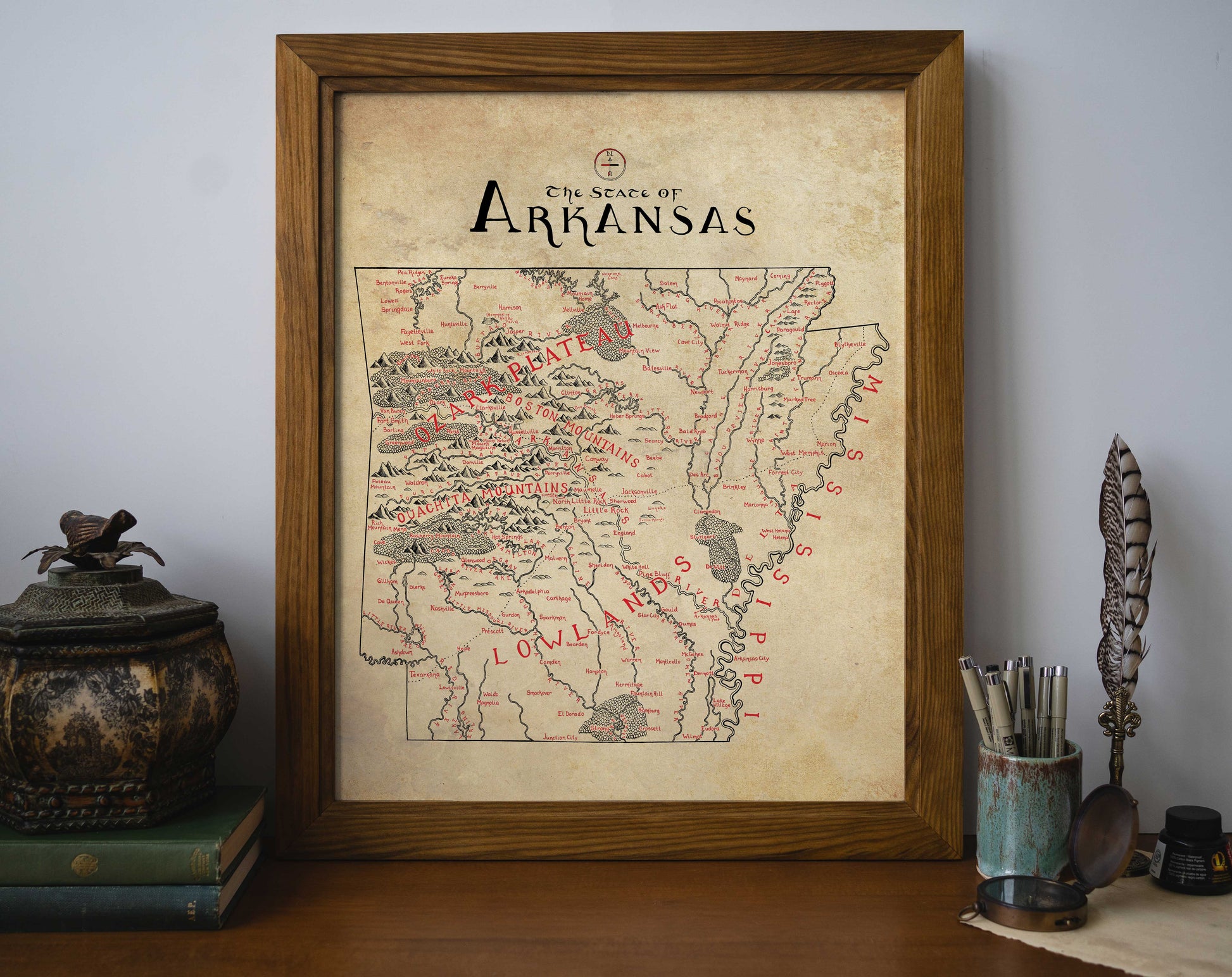 The Arkansas Map by Lord of Maps is a framed hand-drawn print displayed on a desk against a white wall, surrounded by books, a decorative box, potted plant, and pen holder.