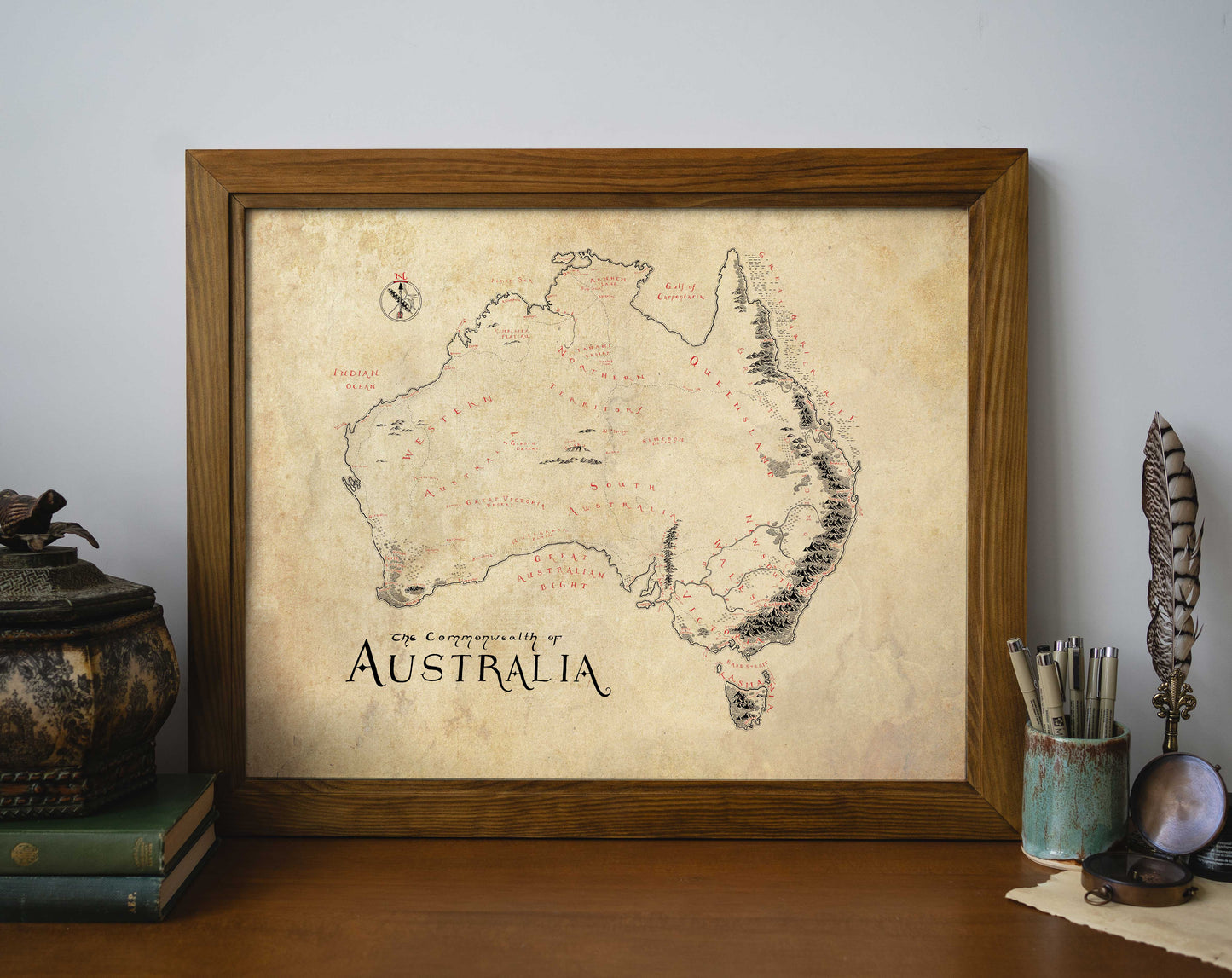 The Australia Map by Lord of Maps is a high-quality print with handwritten labels and a weathered, parchment-style look, shown in a wooden frame on a table alongside books, a quill, and a decorative container.