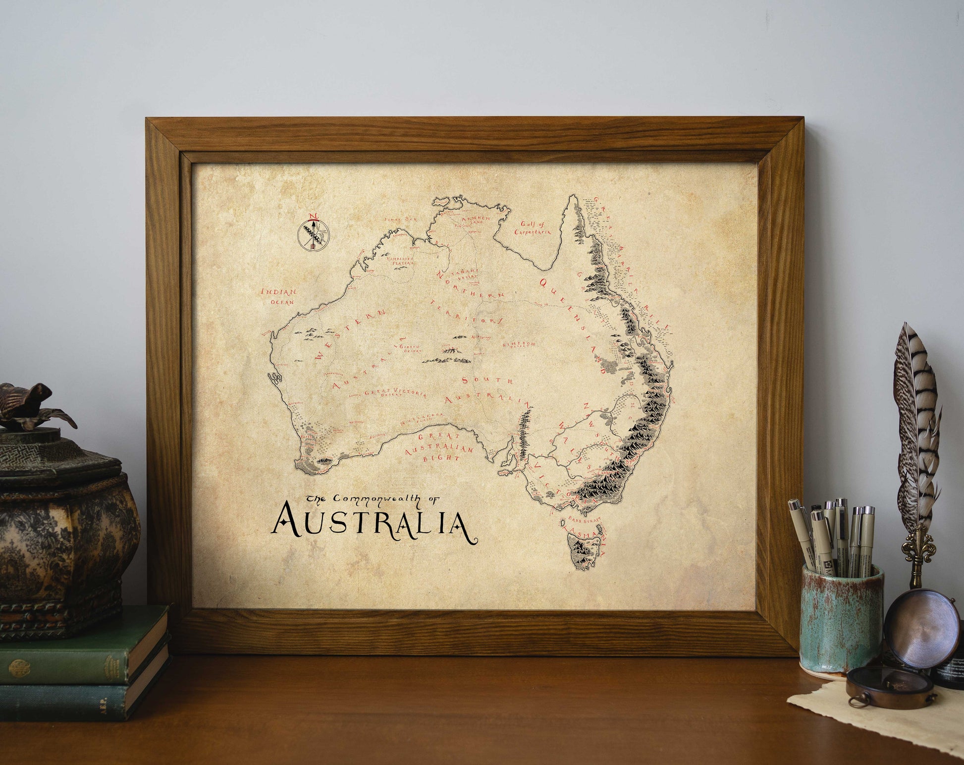The Australia Map by Lord of Maps is a high-quality print with handwritten labels and a weathered, parchment-style look, shown in a wooden frame on a table alongside books, a quill, and a decorative container.