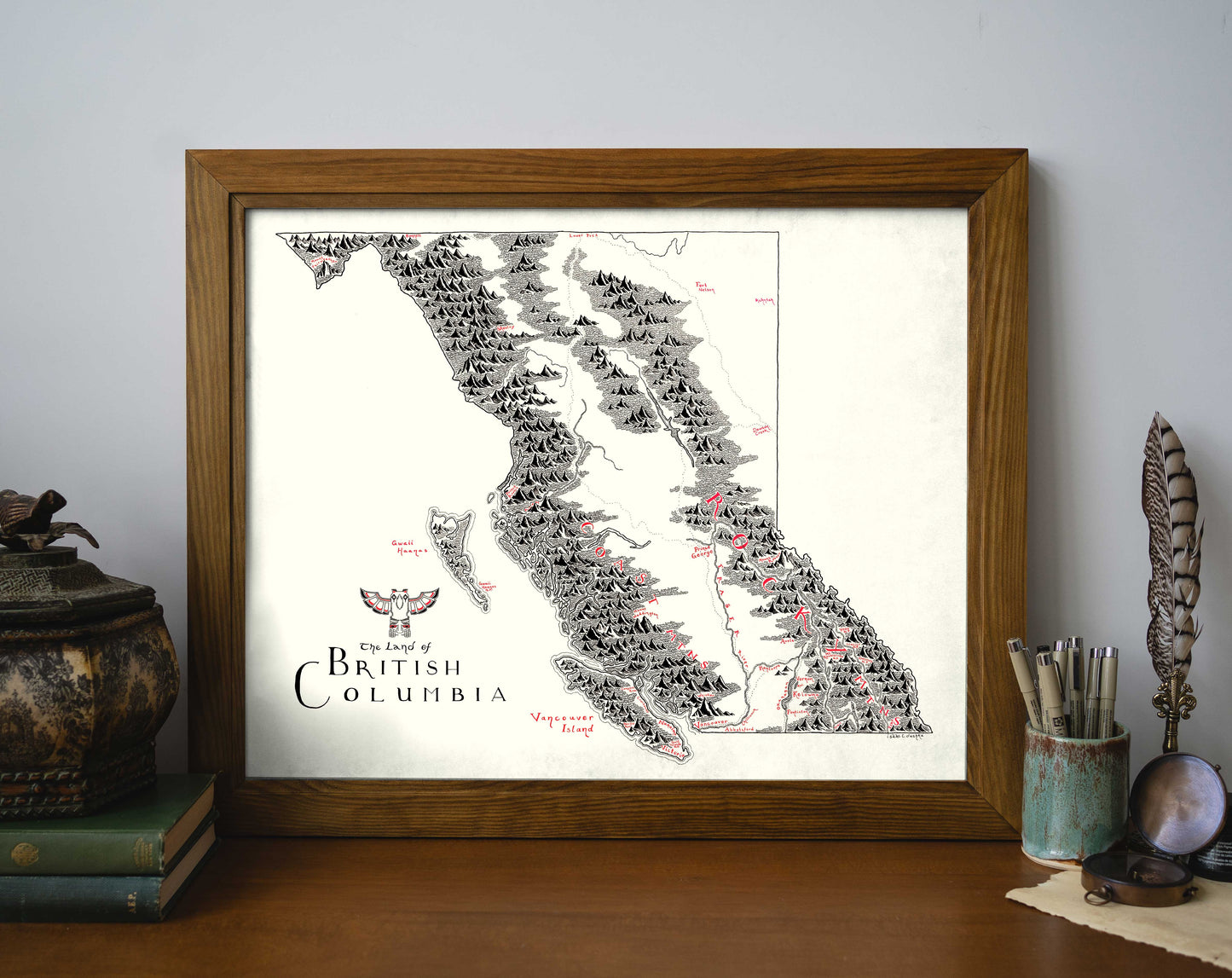 The Lord of Maps British Columbia Map, a hand-drawn framed print, displays mountains and islands with bold black and red labels. It sits on a wooden desk next to books and jars holding pens, feathers, and a paintbrush.