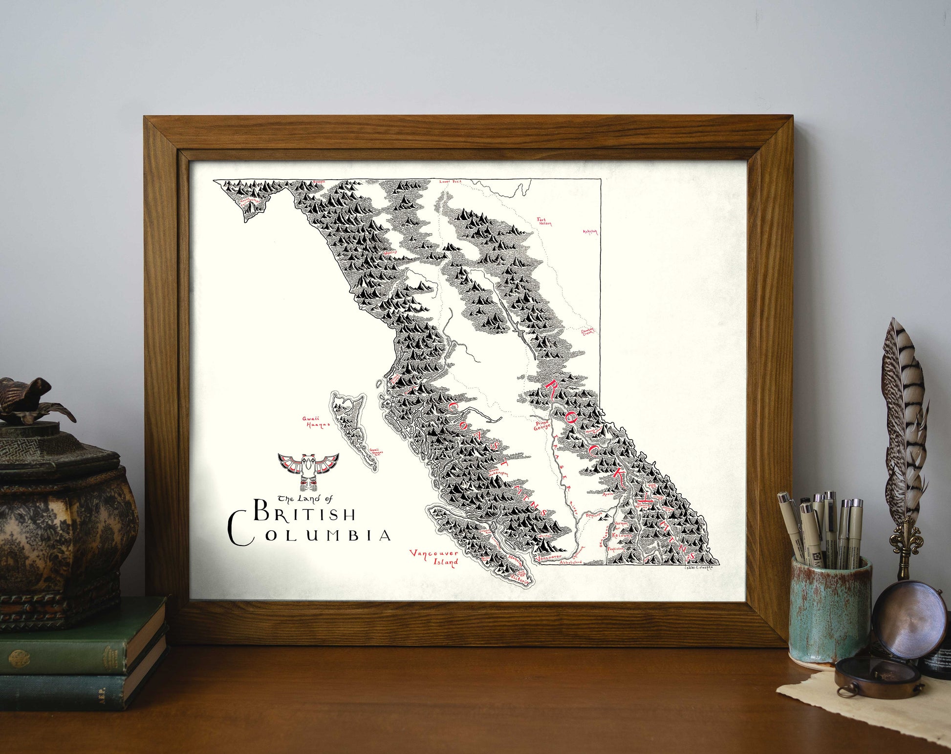 The Lord of Maps British Columbia Map, a hand-drawn framed print, displays mountains and islands with bold black and red labels. It sits on a wooden desk next to books and jars holding pens, feathers, and a paintbrush.