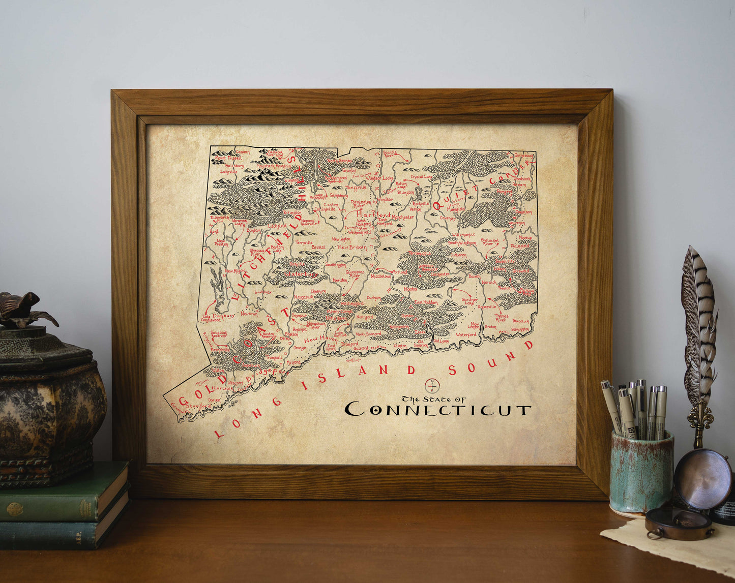 The Lord of Maps Connecticut Map, a vintage-style, hand-drawn print highlighting local features and Long Island Sound, rests framed on a wooden desk amid books, a decorative box, feather quill, and pens.