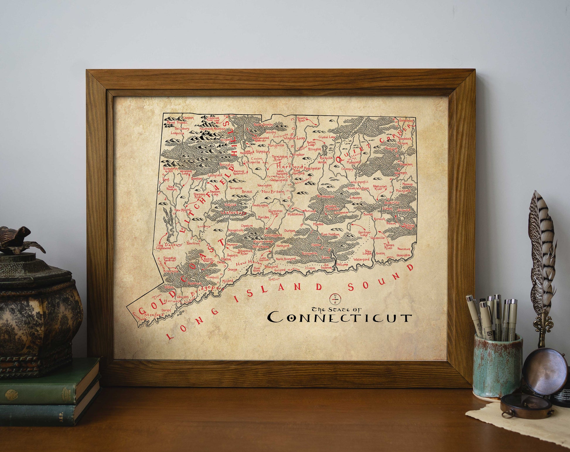 The Lord of Maps Connecticut Map, a vintage-style, hand-drawn print highlighting local features and Long Island Sound, rests framed on a wooden desk amid books, a decorative box, feather quill, and pens.
