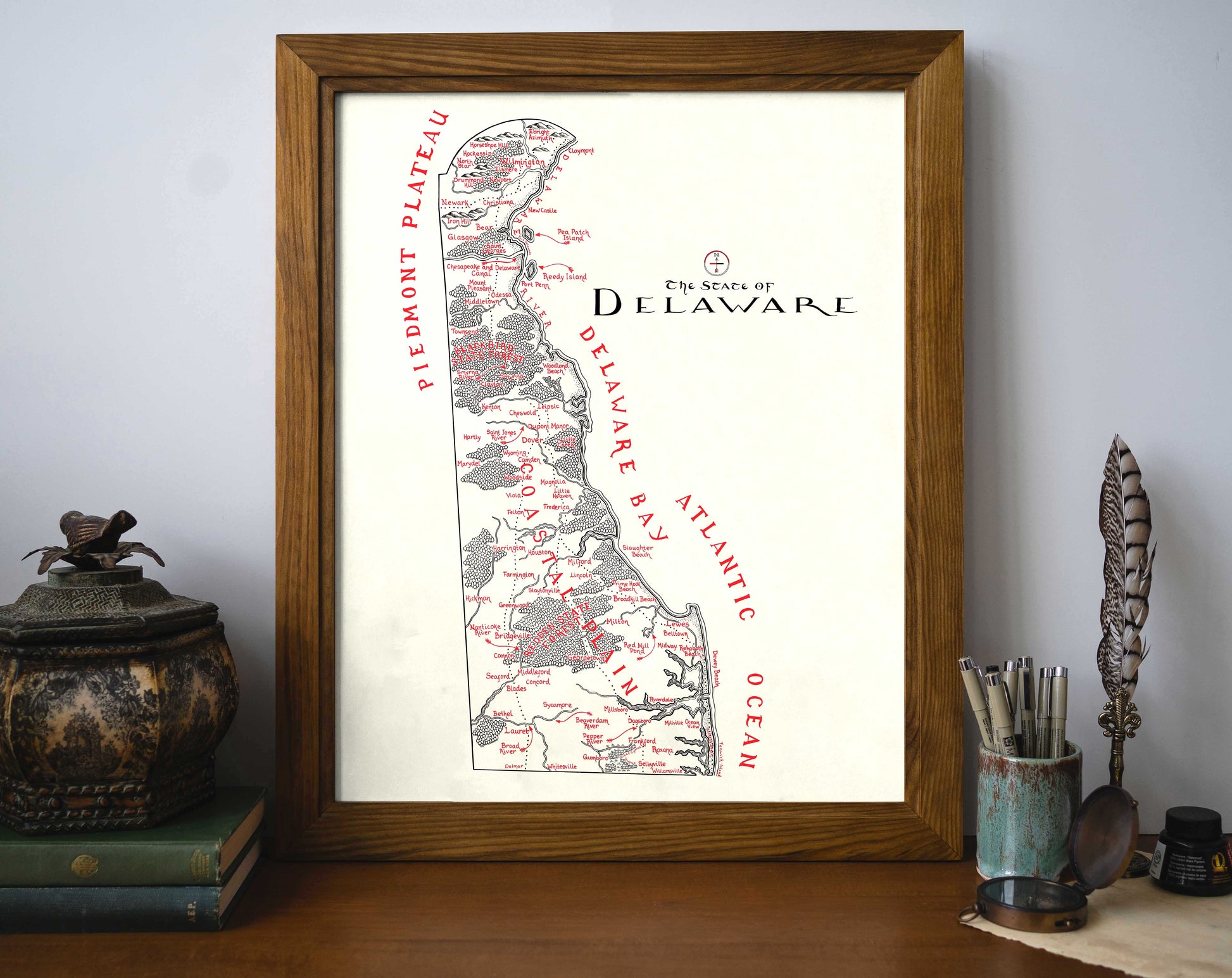 The Delaware Map by Lord of Maps is a framed, hand-drawn print featuring labeled regions and towns, displayed on a wooden desk with decor, notebook, candle, and containers for pens and brushes.