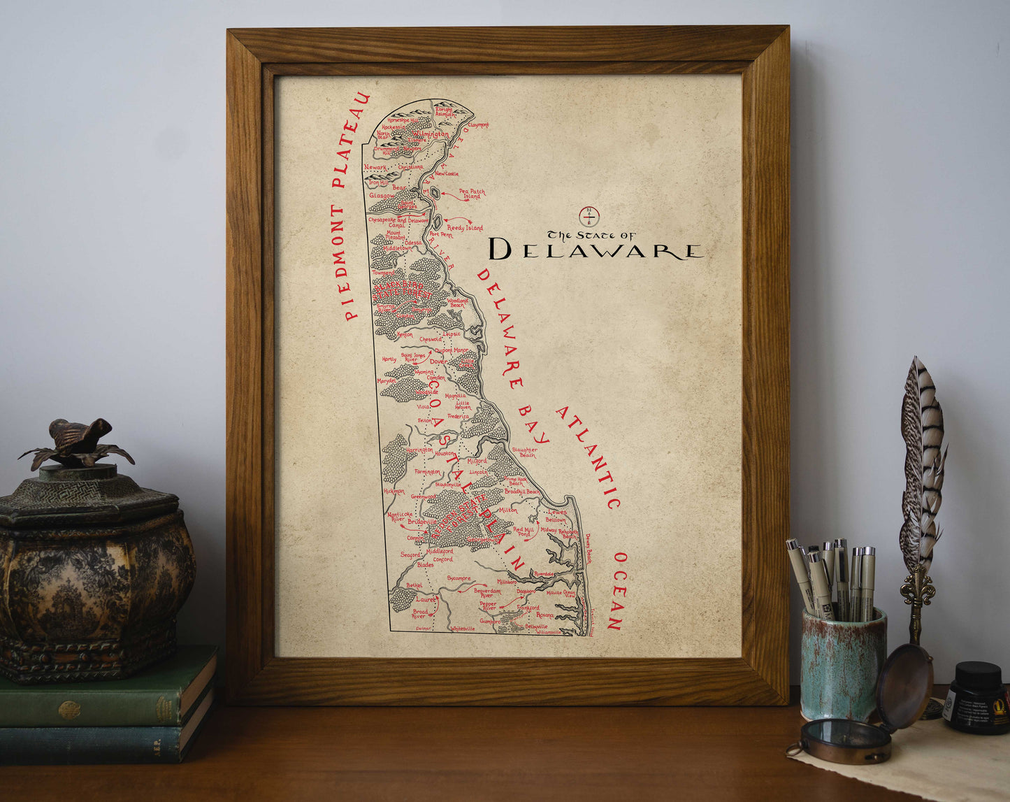 The Lord of Maps Delaware Map, a framed hand-drawn print with labeled regions and cities, stands on a wooden desk beside a decorative box, books, a feather quill, and a cup filled with pens and pencils.