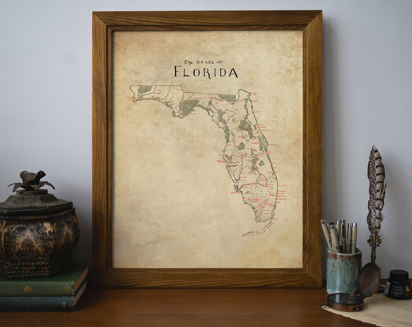 The Lord of Maps Florida Map print, framed in a vintage style, rests on a wooden desk beside a decorative container, stacked books, quill, and pen cup. The plain light wall showcases this unique wall art centerpiece.