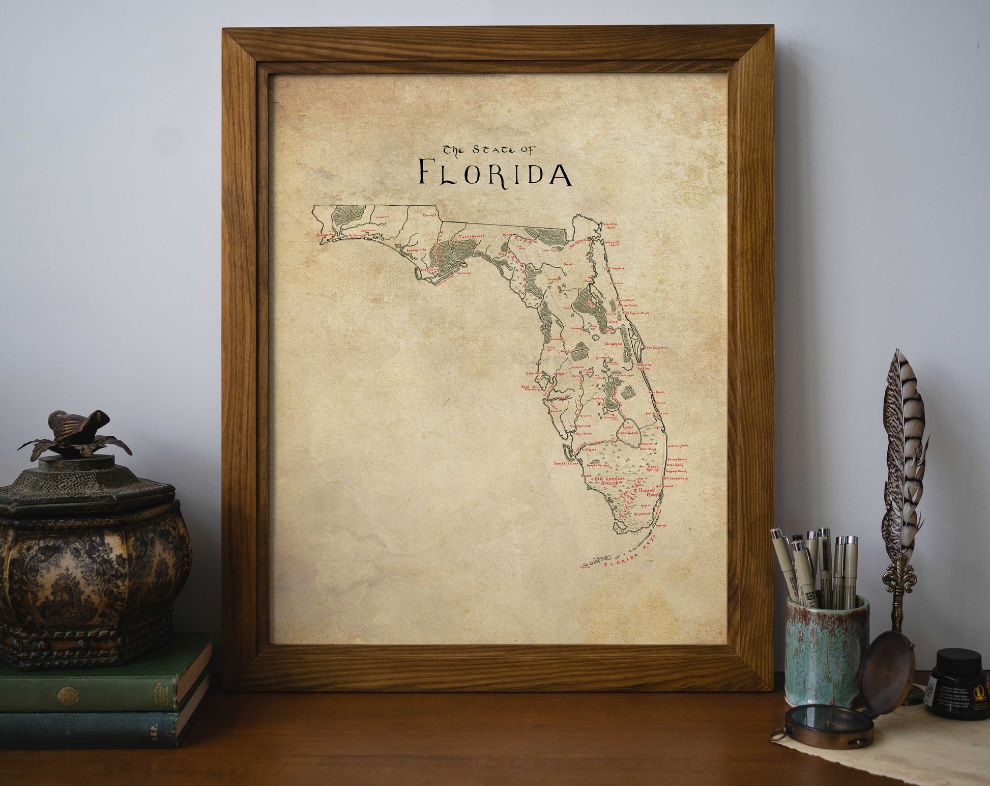 The Lord of Maps Florida Map print, framed in a vintage style, rests on a wooden desk beside a decorative container, stacked books, quill, and pen cup. The plain light wall showcases this unique wall art centerpiece.