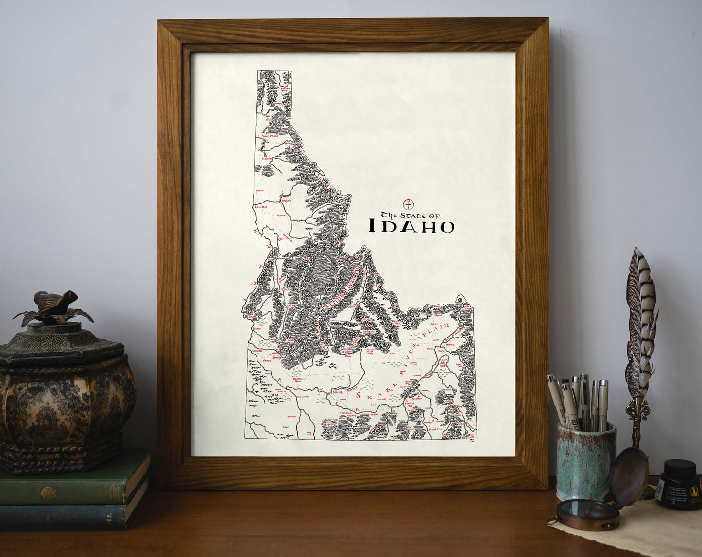 The Lord of Maps Idaho Map, a framed vintage-style hand-drawn print titled "In Search of Idaho," features detailed topography and sits on a desk with books, a decorative jar, quill, and writing utensils.