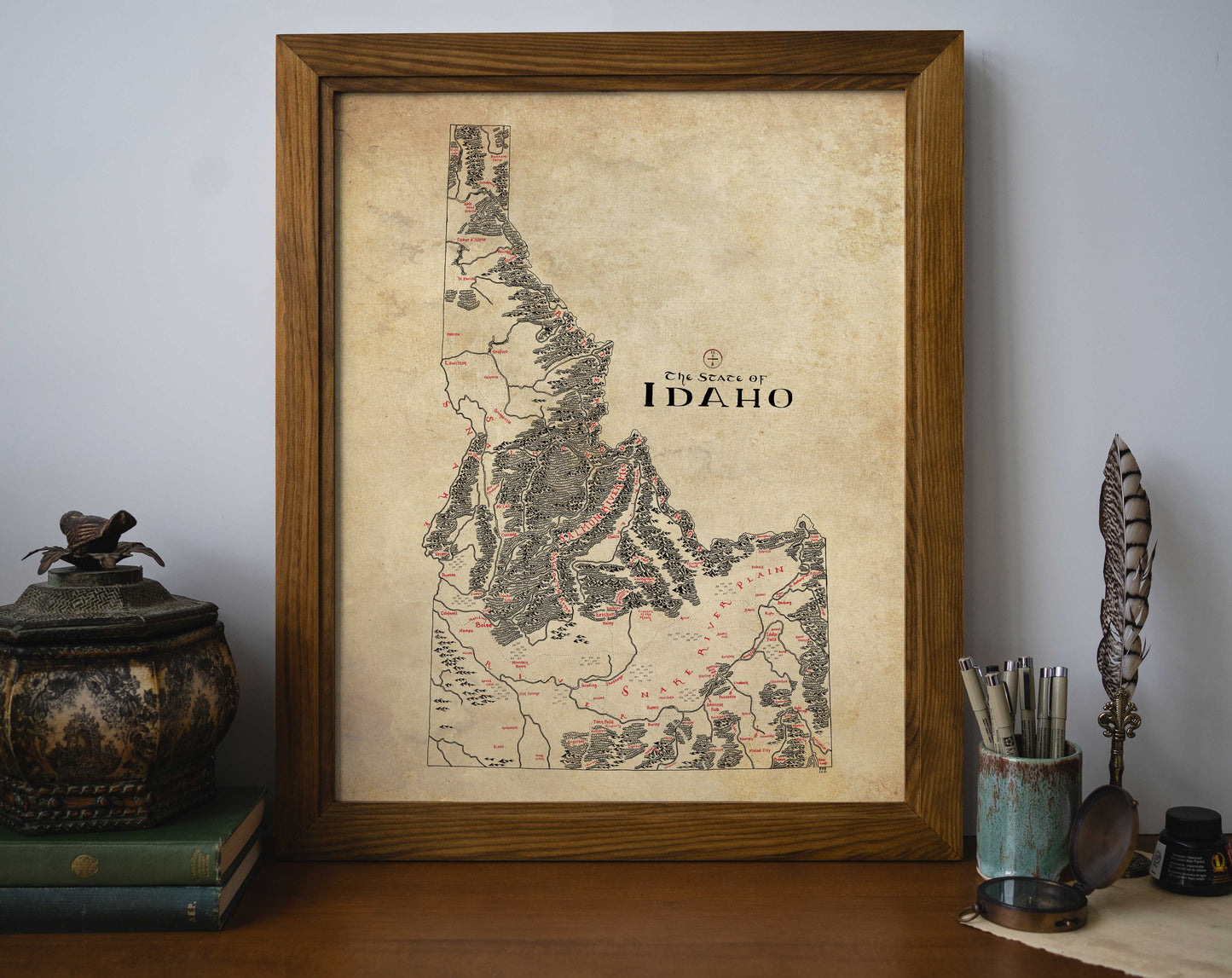 The Idaho Map by Lord of Maps, a vintage-style framed wall art, stands on a desk amid books, a decorative box, an inkwell with quill, pens, and a dish. This hand-drawn print is labeled "EN STATE OF IDAHO.