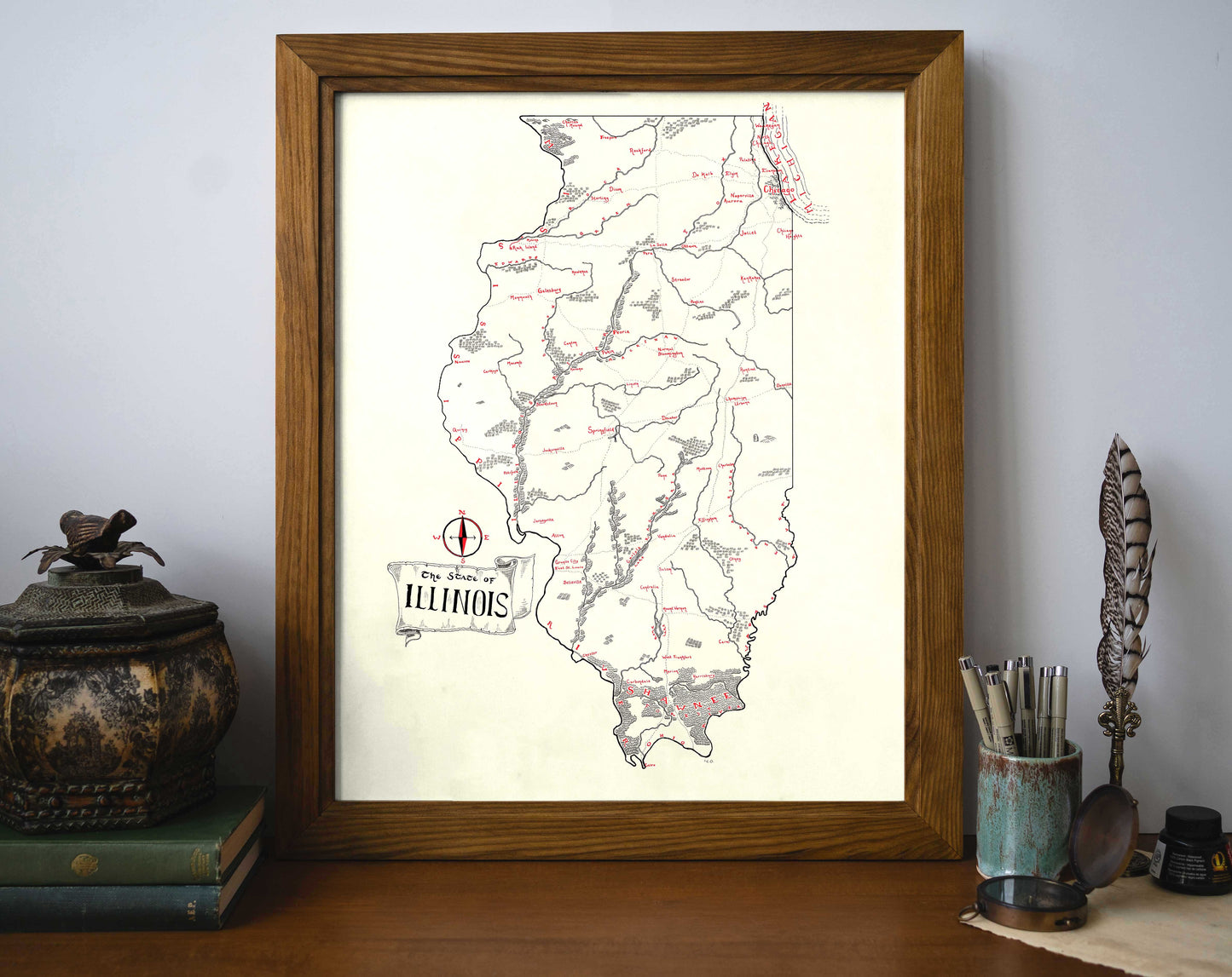 The Illinois Map by Lord of Maps, a framed vintage-style print in black and red ink, features rivers, cities, and towns. Displayed on a desk alongside antique objects, books, a feather quill, and a cup holding pens and pencils.
