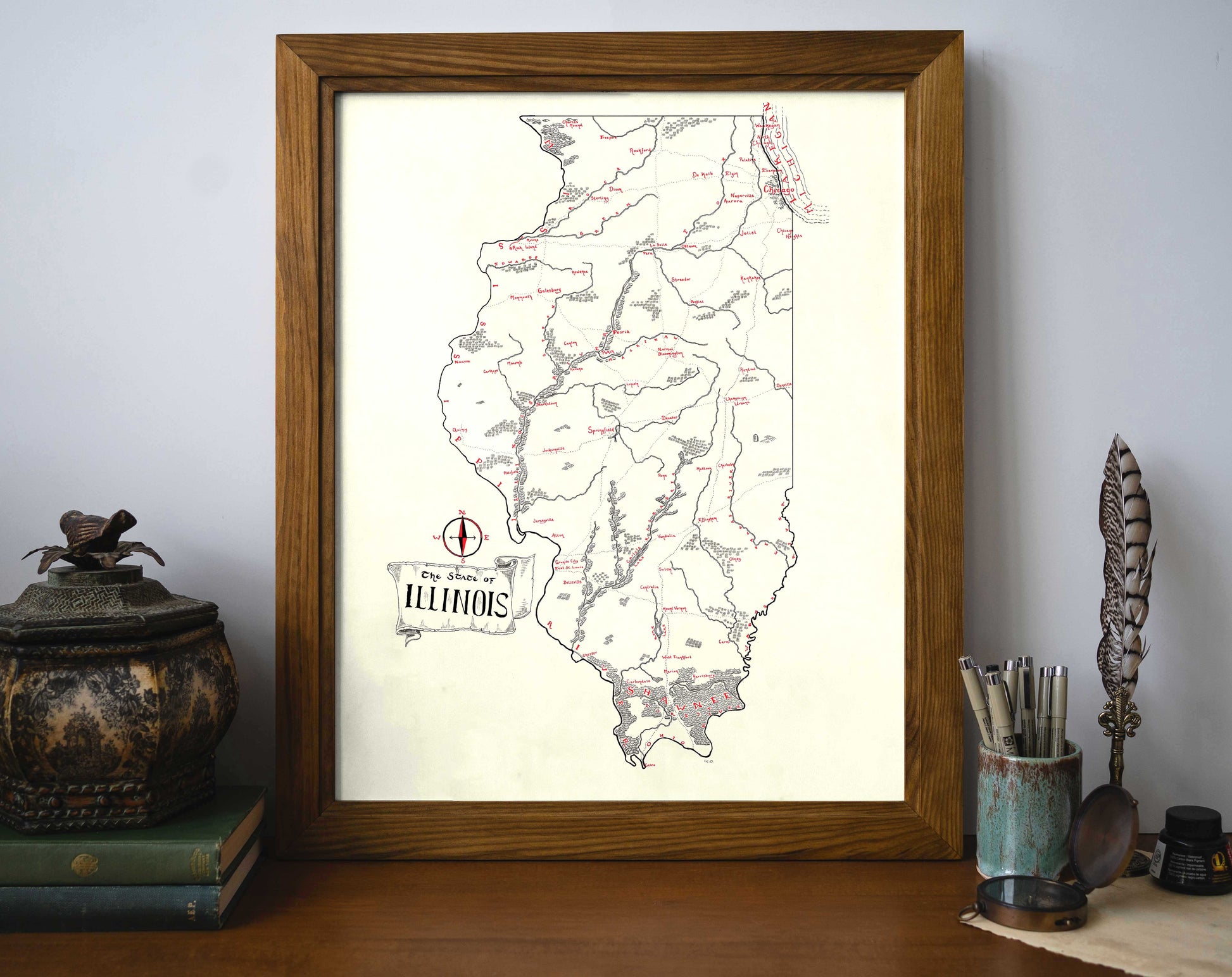 The Illinois Map by Lord of Maps, a framed vintage-style print in black and red ink, features rivers, cities, and towns. Displayed on a desk alongside antique objects, books, a feather quill, and a cup holding pens and pencils.