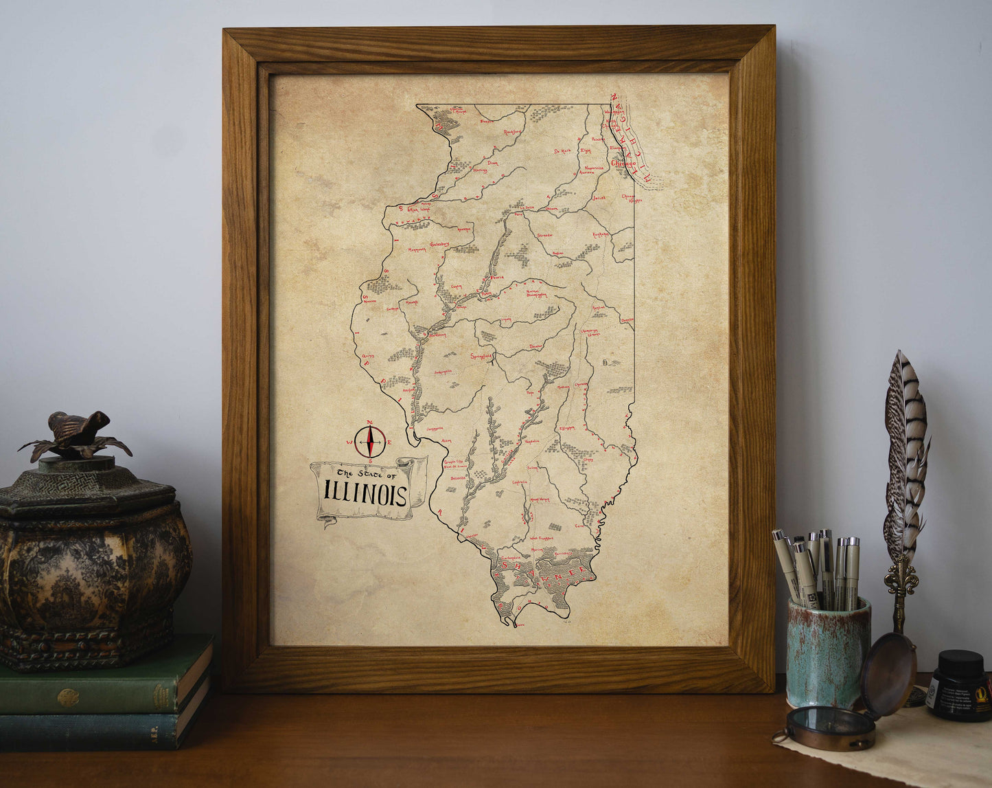 The Illinois Map by Lord of Maps rests framed on a desk with books, an inkwell, a quill, and a cup of pens and scissors. This vintage-style map print highlights rivers and cities in red and black text.