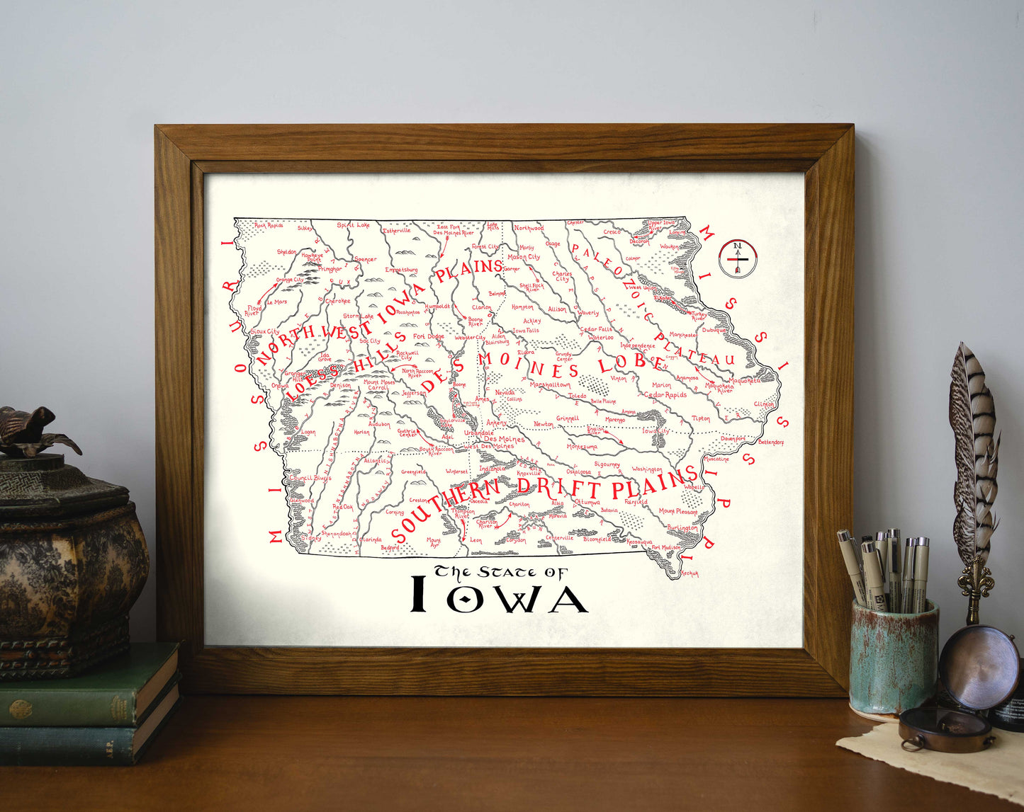 The Lord of Maps "Iowa Map" framed wall art features vintage-style detailing with regional names and cities in red text. Displayed on a desk, it's complemented by books, a potted plant, and a decorative feather stand.