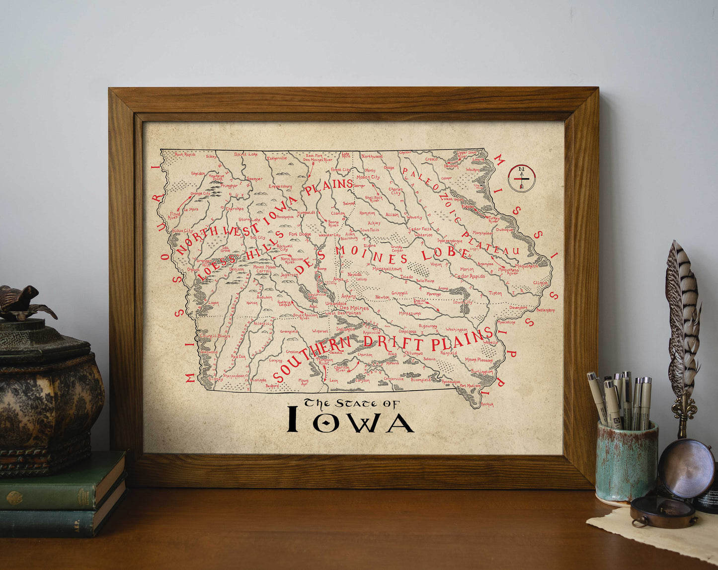 The Lord of Maps Iowa Map is a framed vintage wall art print featuring regions such as North Ilion Plains, Des Moines Lobe, and Southern Drift Plains, shown on a wooden shelf with books, a vase, and writing utensils.