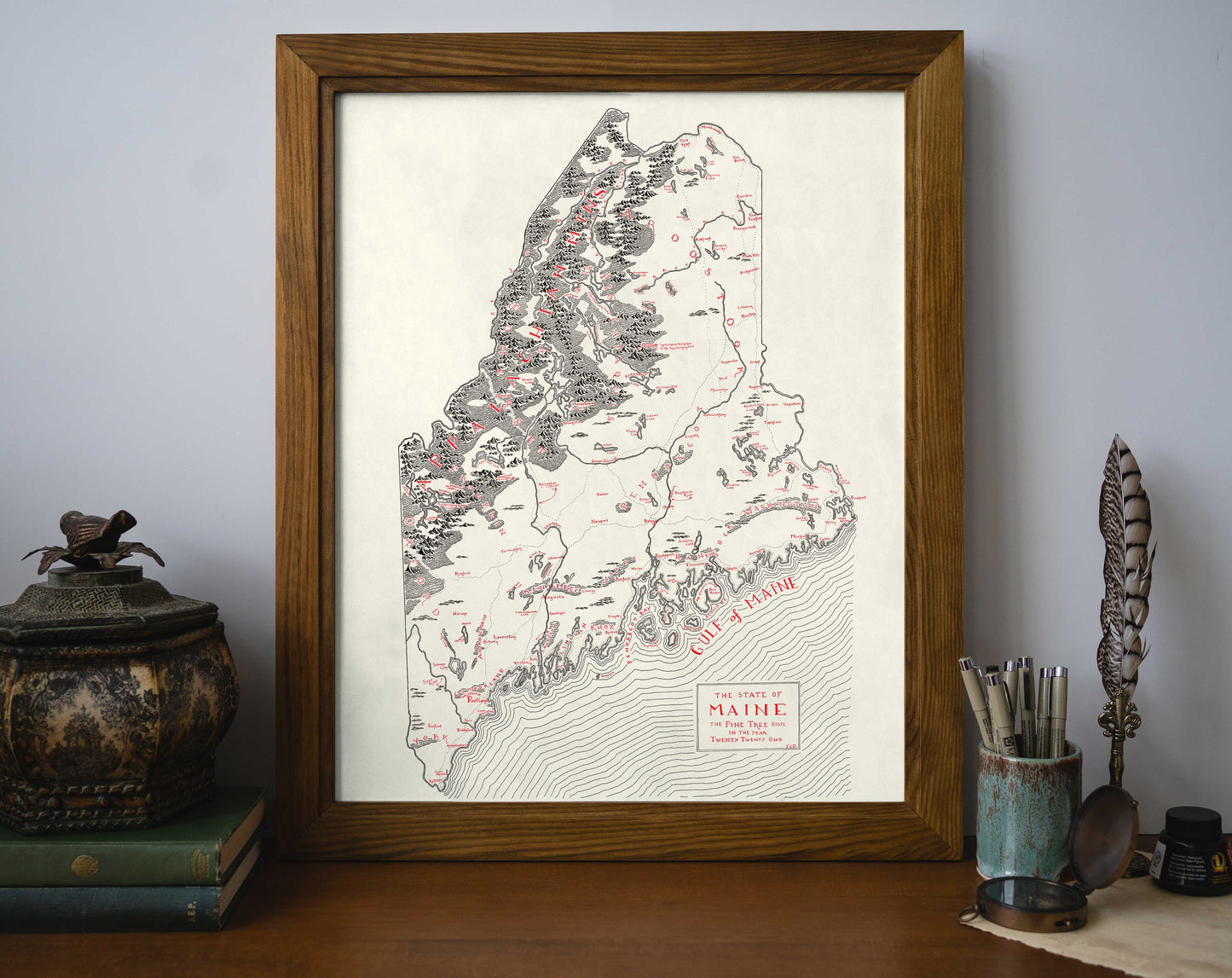 The Maine Map by Lord of Maps is a framed black and white hand-drawn print featuring red accents to highlight select locations, displayed on a desk with books, a decorative container, pens, and a feather for added style.