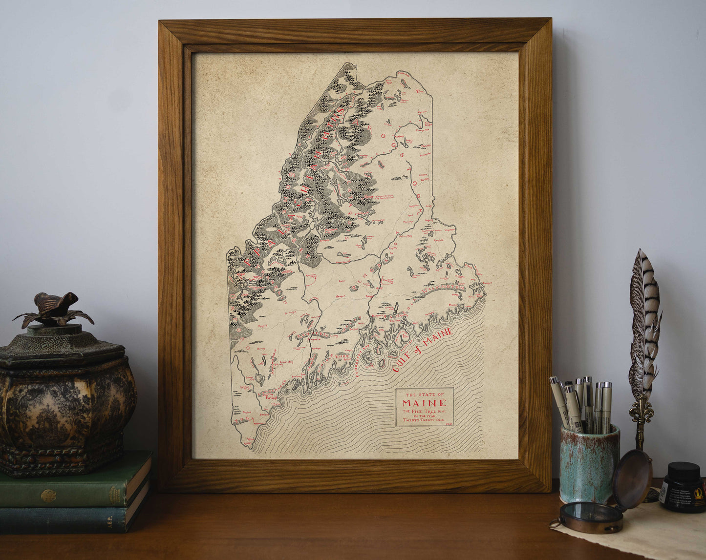 The Lord of Maps Maine Map, a high-quality framed print, shows vintage-style topography and labeled towns in black and red, perfect for displaying on your desk with books and writing accessories.