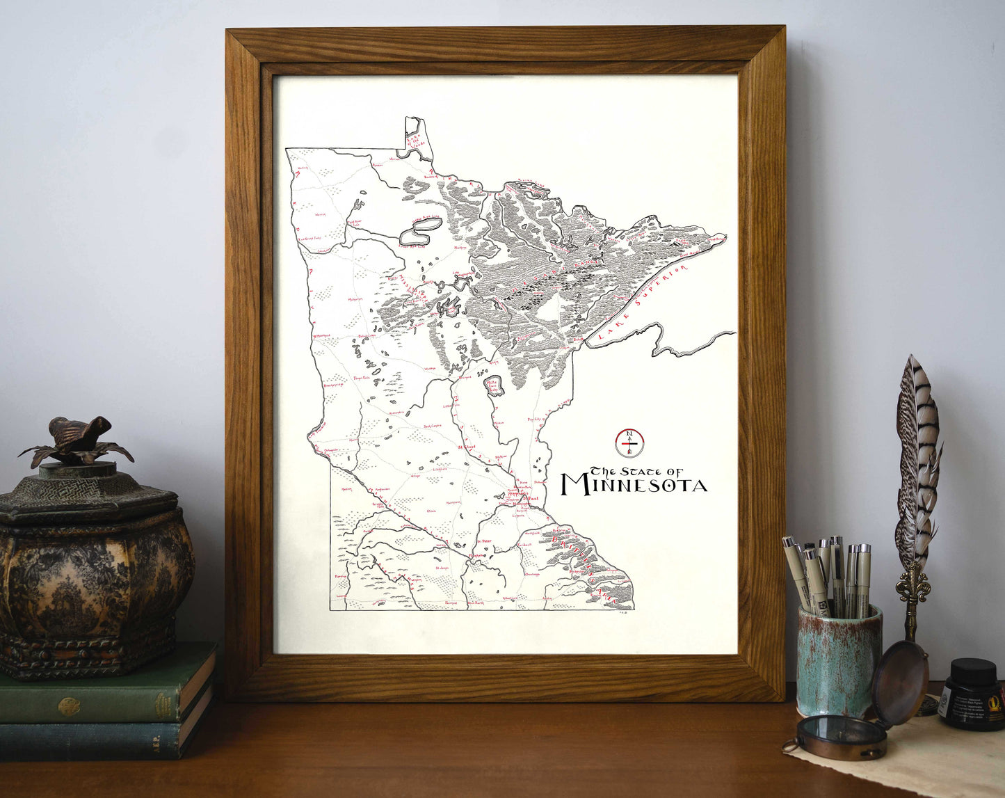 The Lord of Maps Minnesota Map, a framed vintage-style print with detailed markings and text, sits on a desk against a wall, surrounded by books, a decorative jar, feather quill, and cups with pens and pencils.