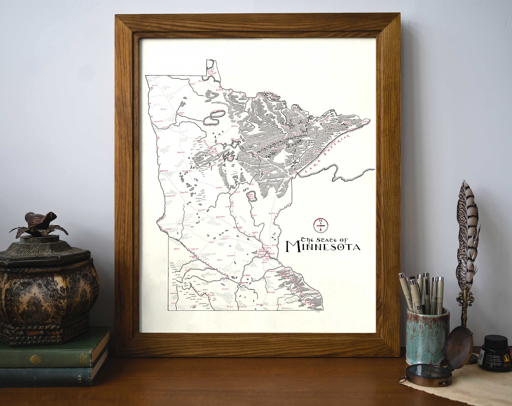 The Lord of Maps Minnesota Map, a framed vintage-style print with detailed markings and text, sits on a desk against a wall, surrounded by books, a decorative jar, feather quill, and cups with pens and pencils.