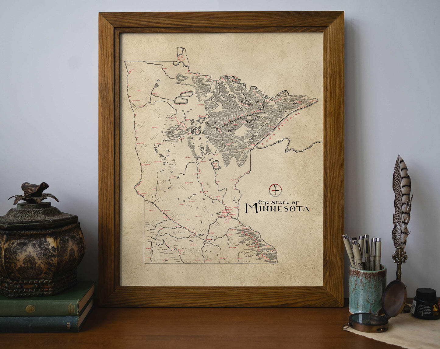The Lord of Maps Minnesota Map, beautifully framed, rests on a table beside stacked books, a decorative container, and writing tools, adding a cozy, vintage touch to your space with this high-quality map art.